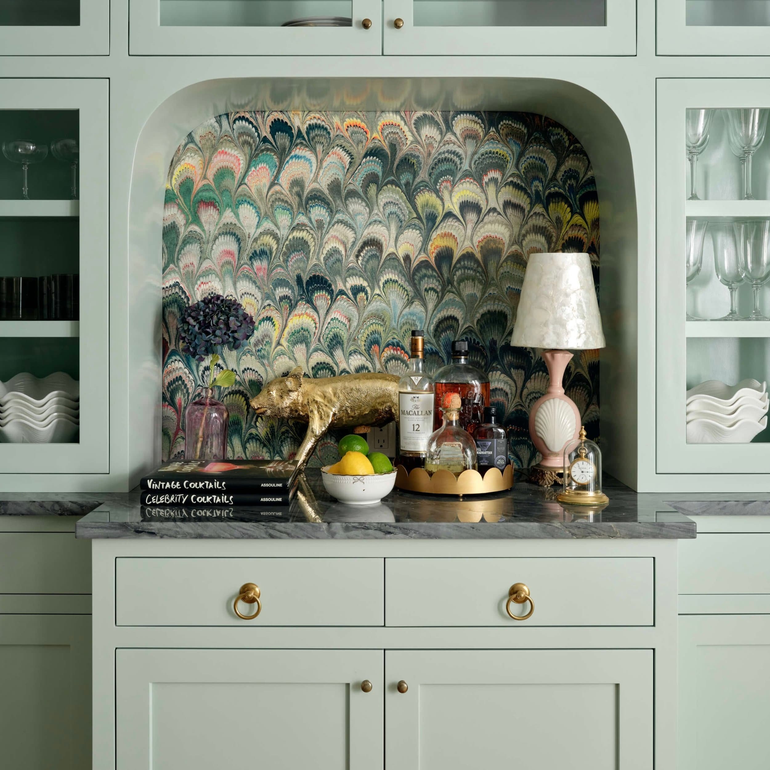 Decorative kitchen countertop with a collection of liquor bottles, a white bowl with lemons and limes, a gold animal sculpture, a pink and white table lamp, a small clock, and stacked cocktail books, set against a patterned wallpaper and pastel cabin