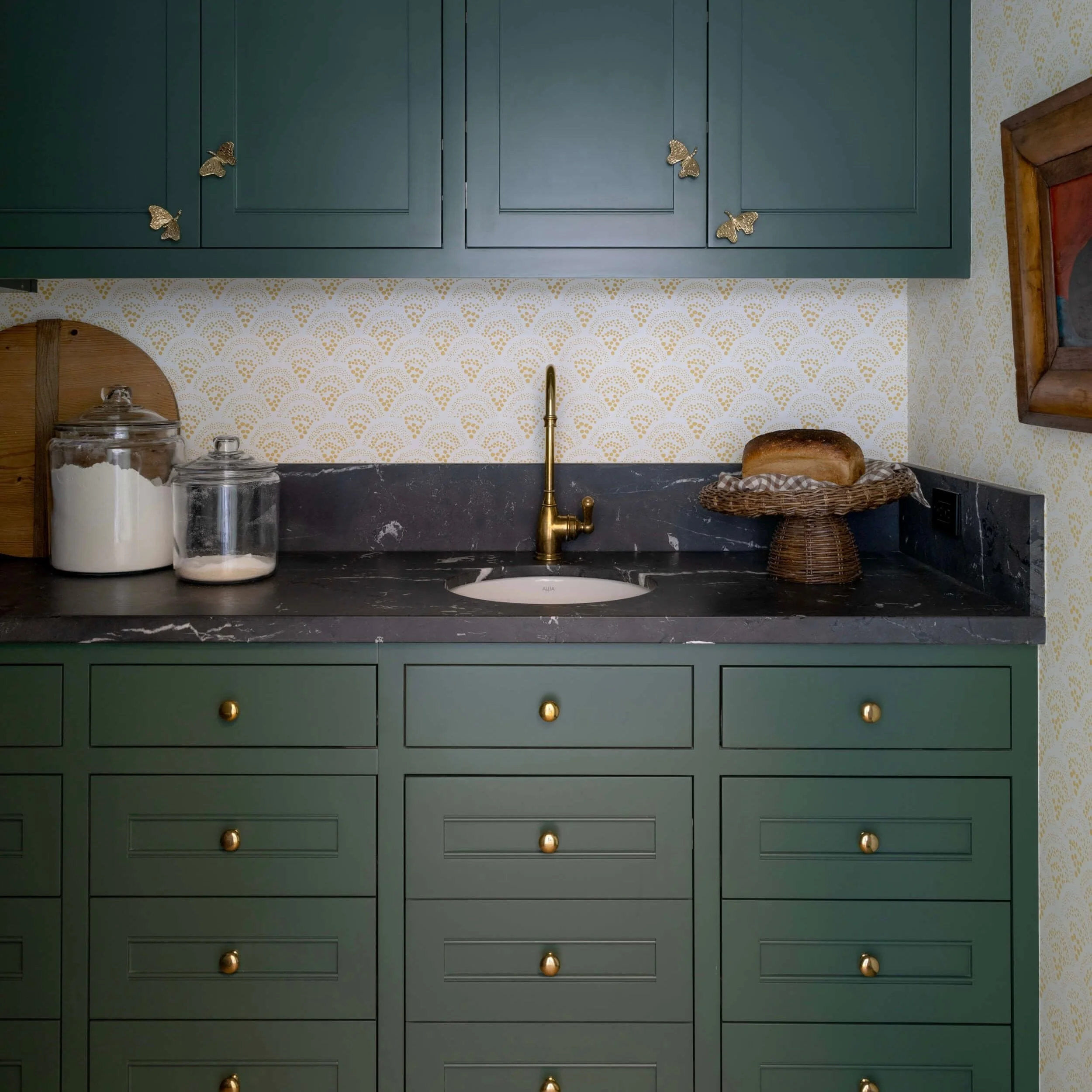 Green kitchen cabinets with gold knobs, window with wooden frame, black marble countertop, gold faucet, paper bag with bread, glass jars with flour and sugar, wallpaper with a gold pattern, and decorative gold butterflies