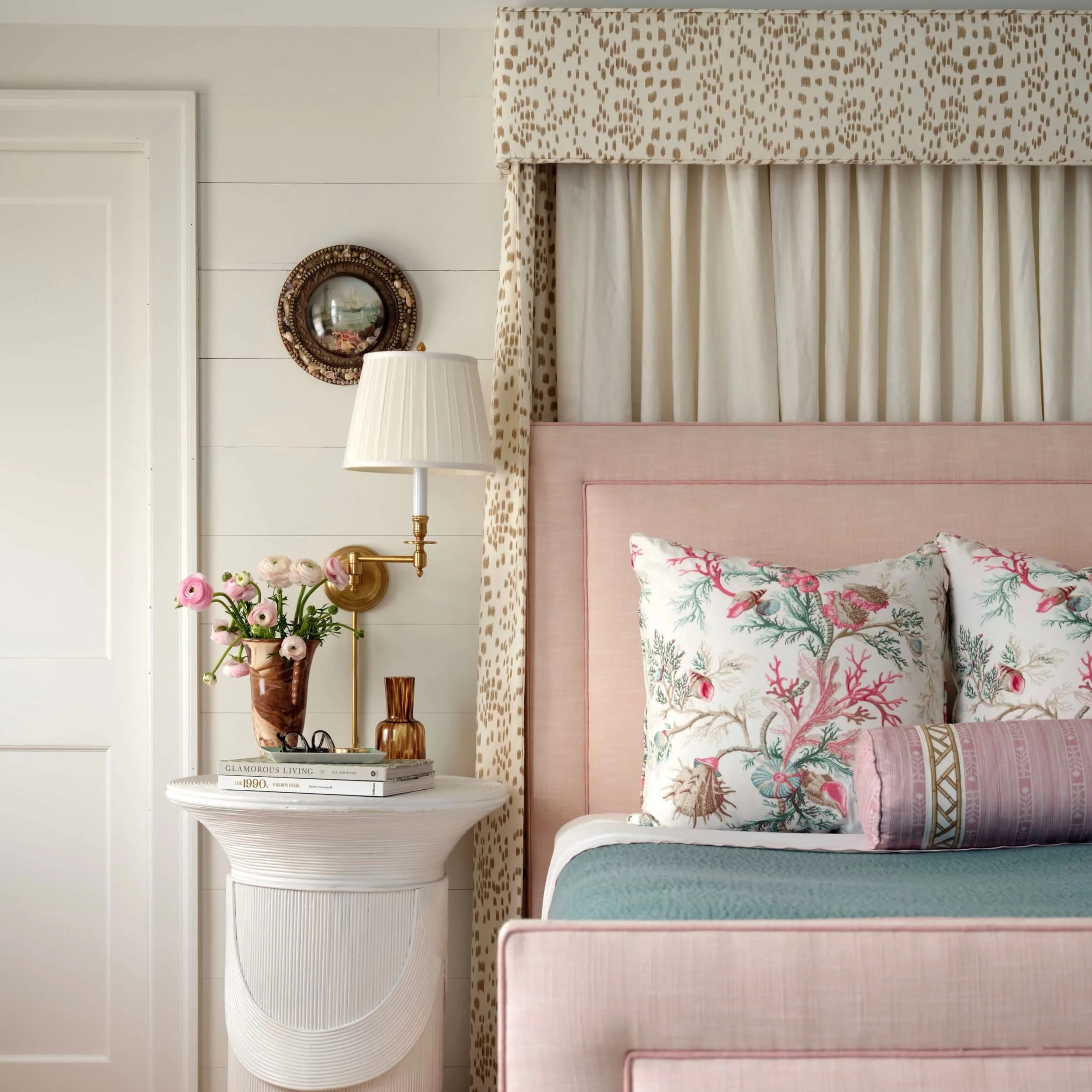 A bedroom with a pink upholstered headboard, bedding with pink and green botanical prints, a nightstand with a vase of pink and white flowers, books, and decorative items, beige patterned curtains, a white wall with a small round mirror, and a wall-m