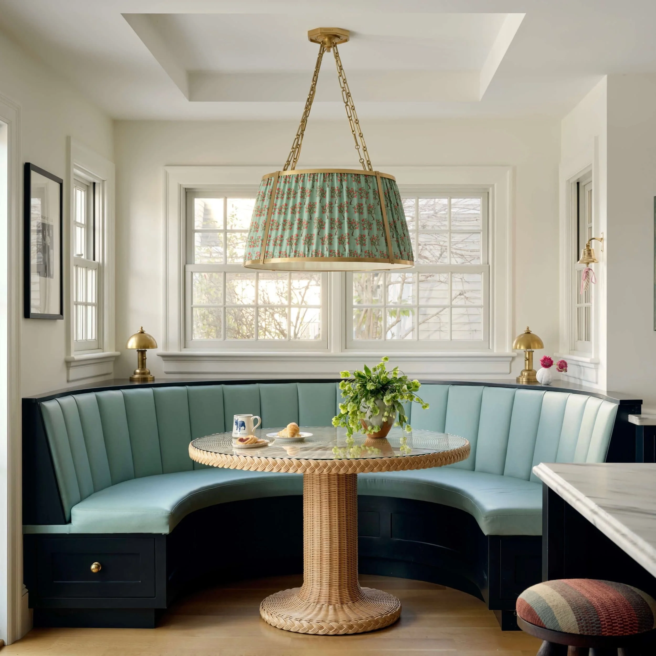 A cozy corner of a dining nook with a curved, mint green upholstered banquette, a round wicker table with a potted plant, two mugs, and pastries, illuminated by natural light from multiple windows with brass lamps on the window sills and a floral lam