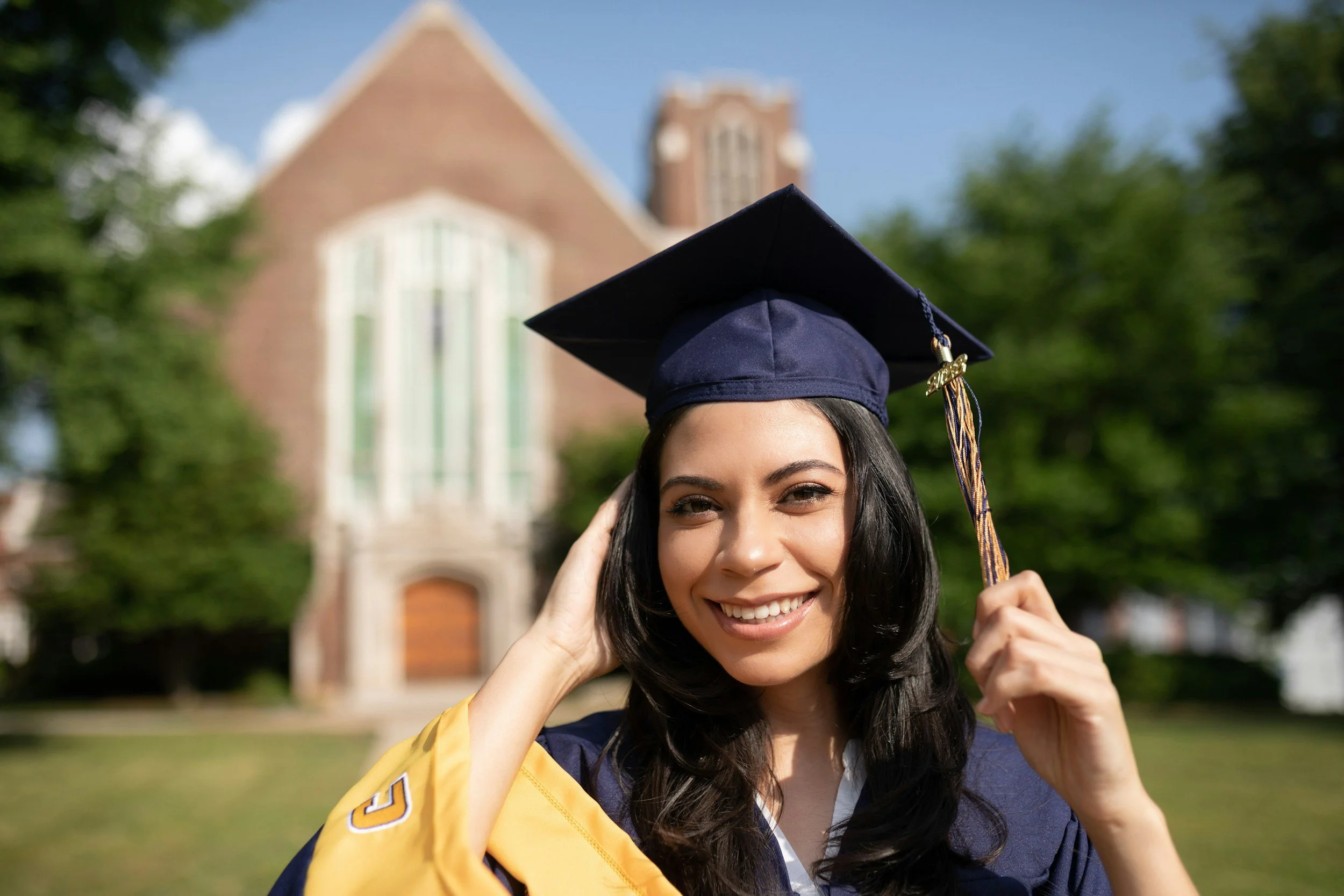 Young woman in cap and gown celebrating graduation outdoors in front of a brick church with green trees and blue sky.