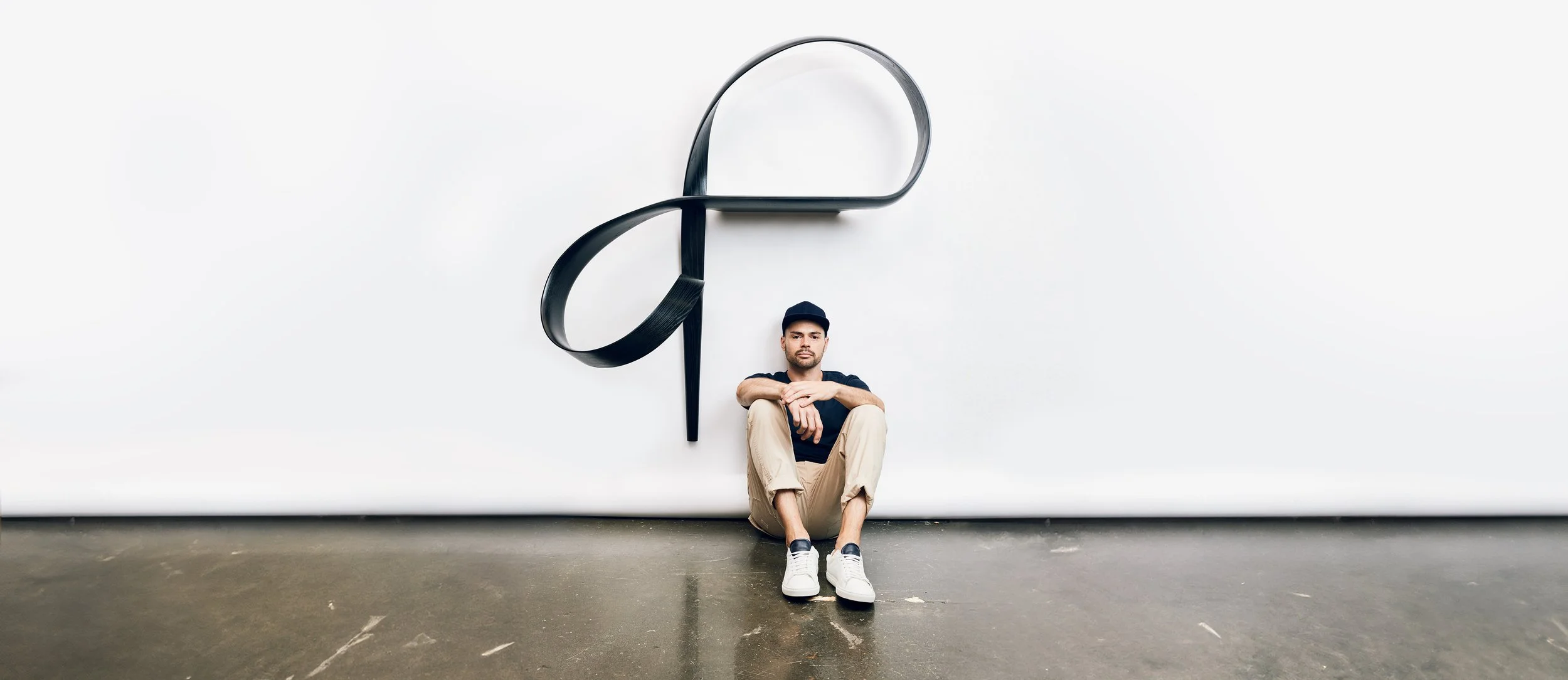 Adam Bentz seated beneath a large bent wood wall sculpture in his studio, highlighting sculptural form, scale, and architectural composition.