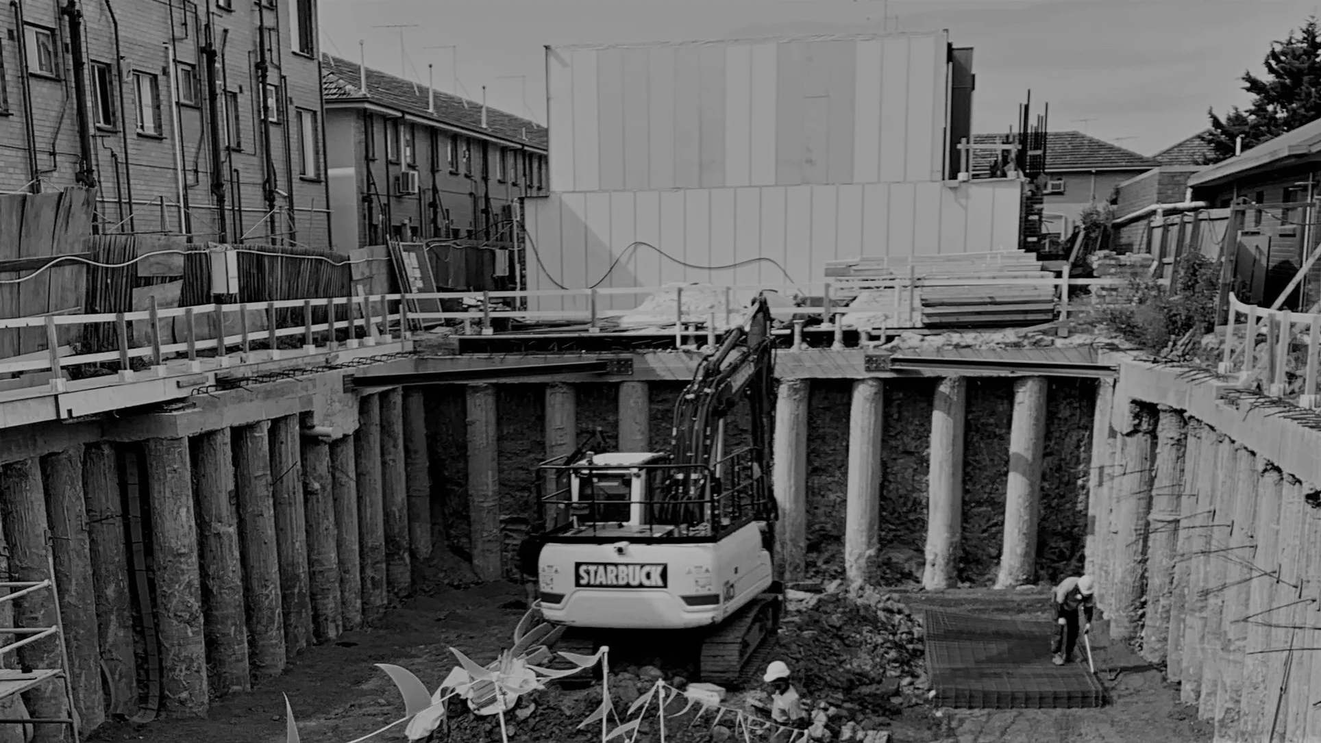 Construction site with a basement excavation, small excavator, wooden seawalls, and construction workers wearing helmets.