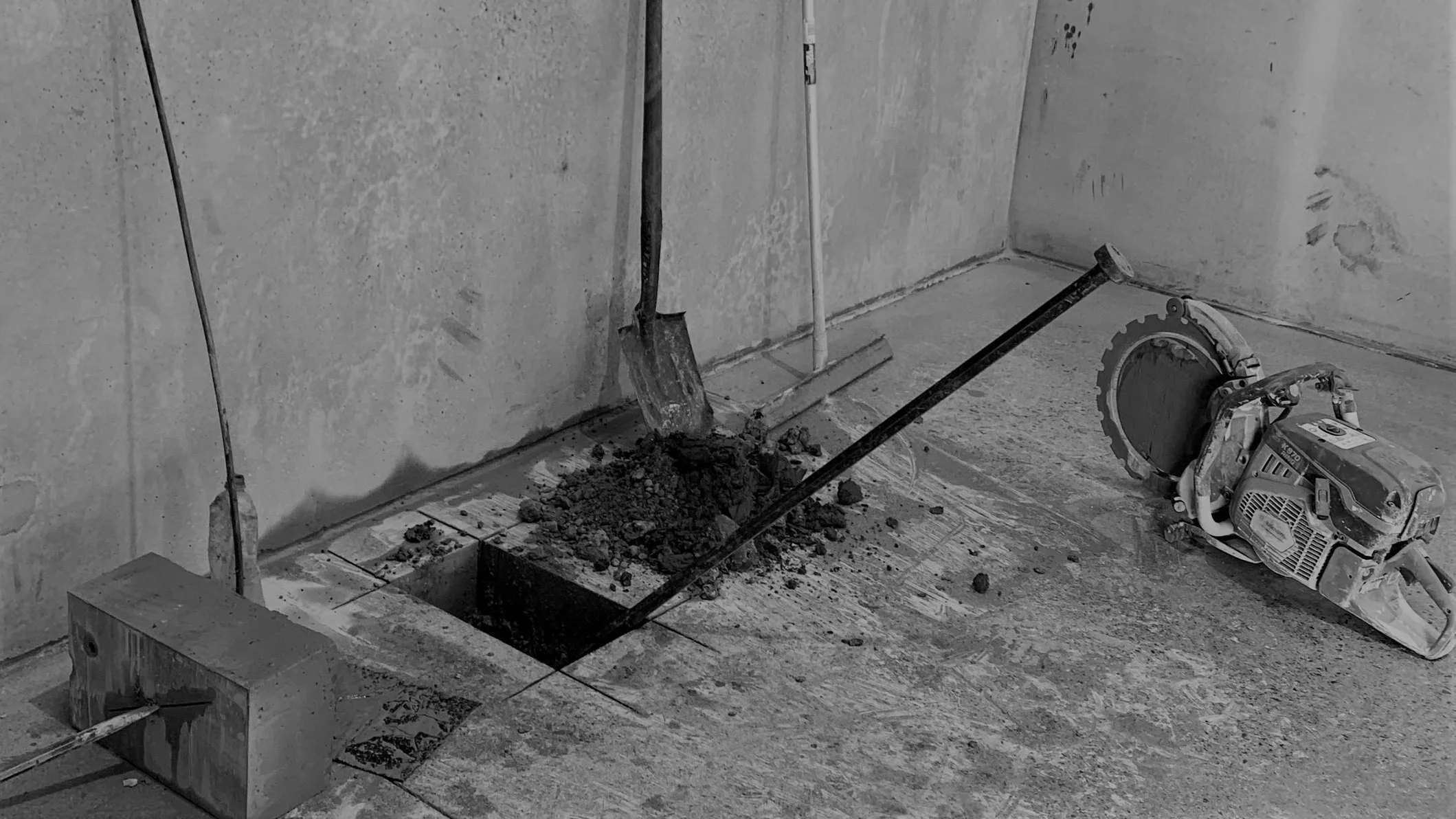 Construction site with tools including a shovel and a concrete saw on a floor with an opening in the ground and exposed concrete walls.