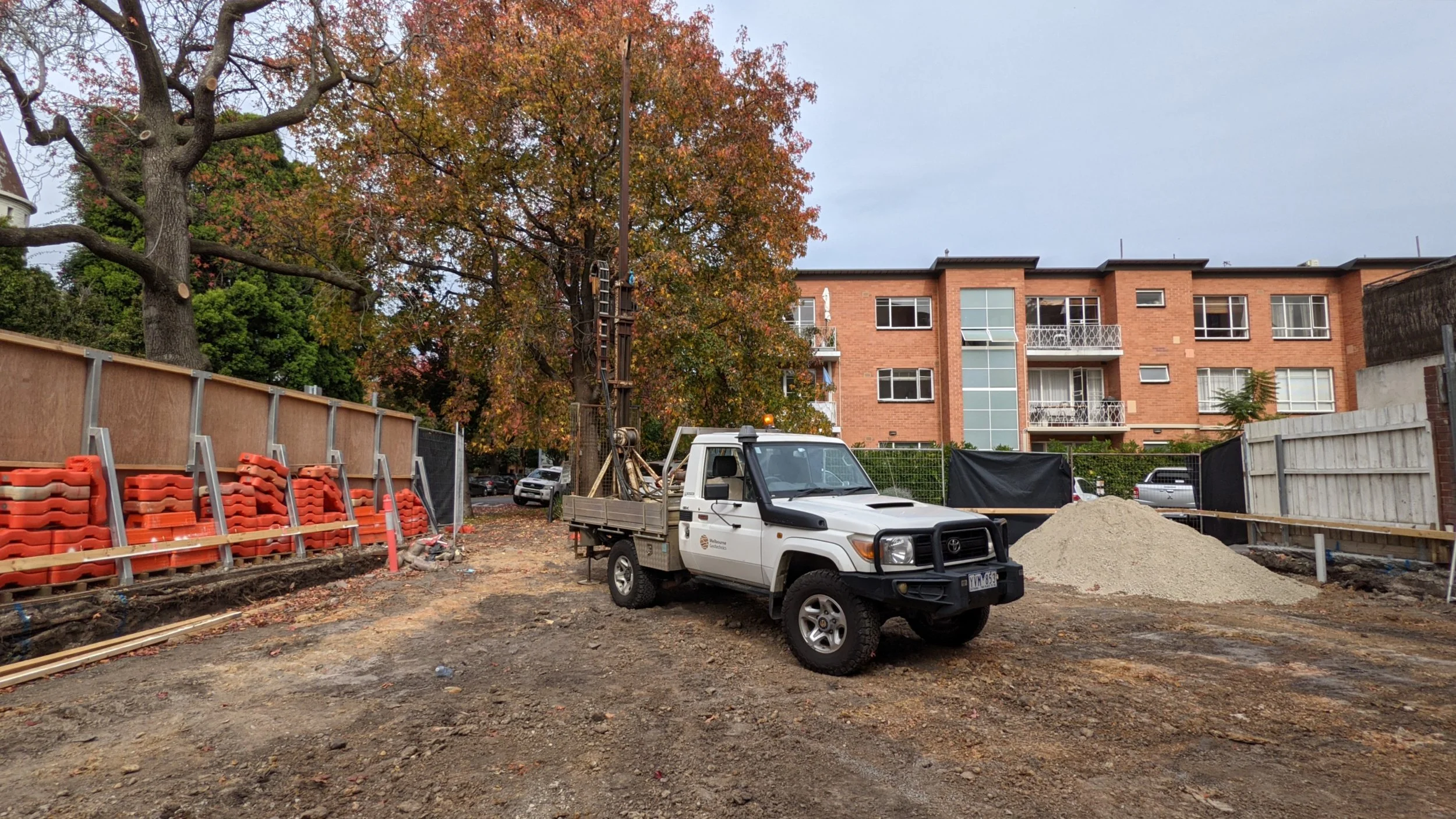 Construction site with a white pickup truck, orange safety barriers, a large tree with autumn leaves, and a three-story brick residential building in the background.