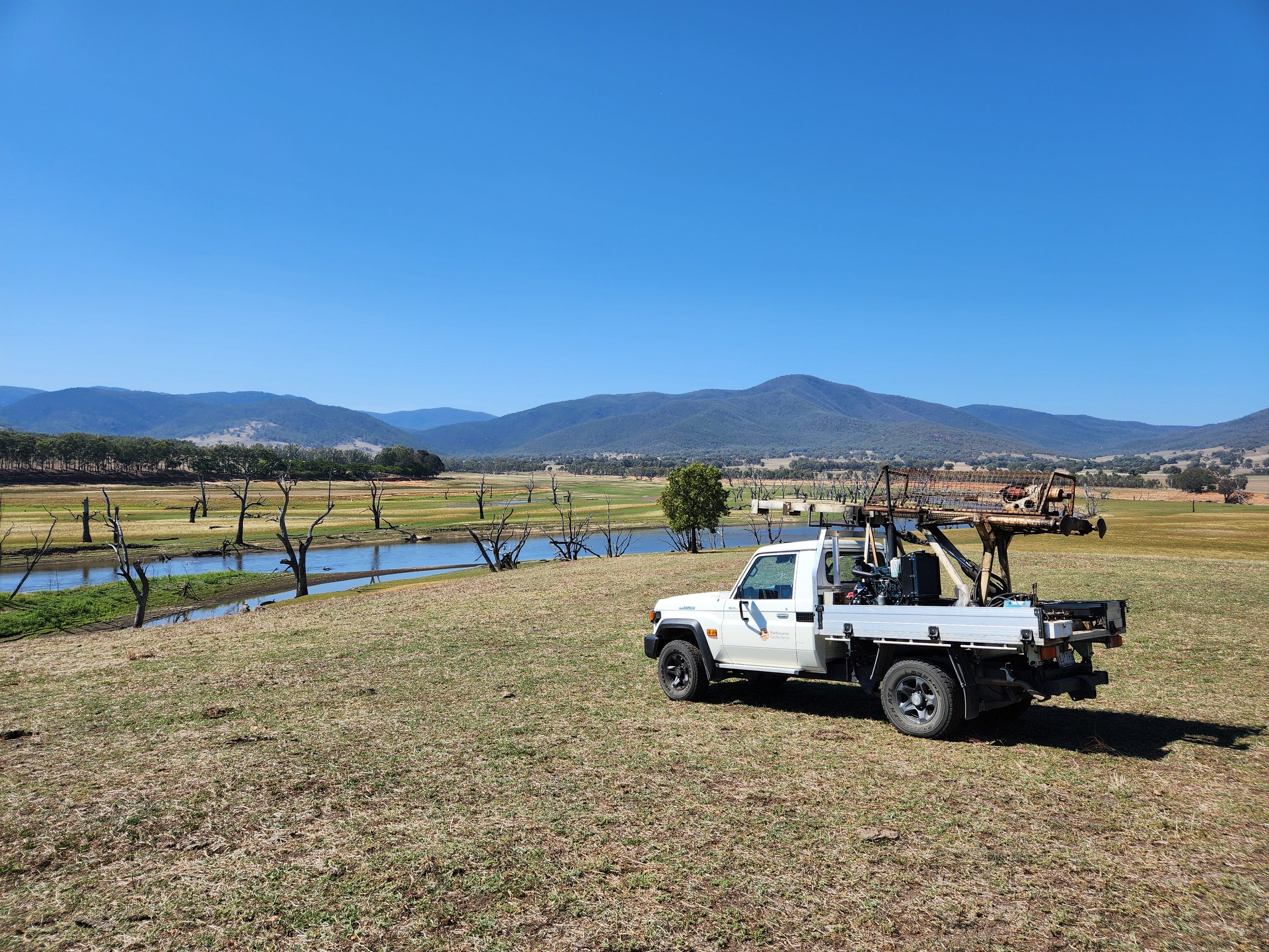 A white utility truck with a rusty ladder attachment parked on a grassy field near a river, with leafless trees, green patches, mountains, and clear blue sky in the background.