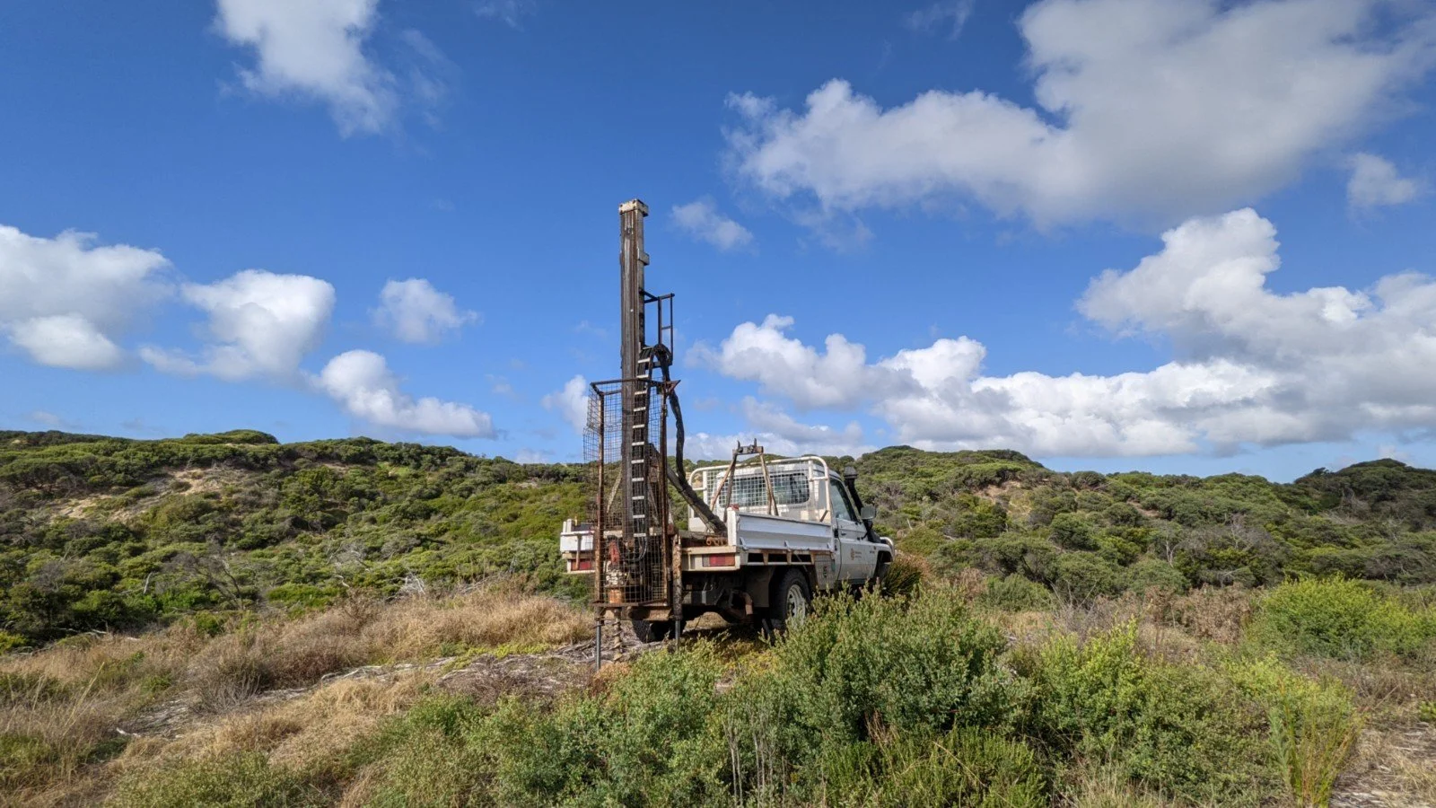 A white utility truck with a tall, drilling rig mounted on its bed, set in a grassy, shrub-covered landscape with rolling hills and a partly cloudy blue sky.