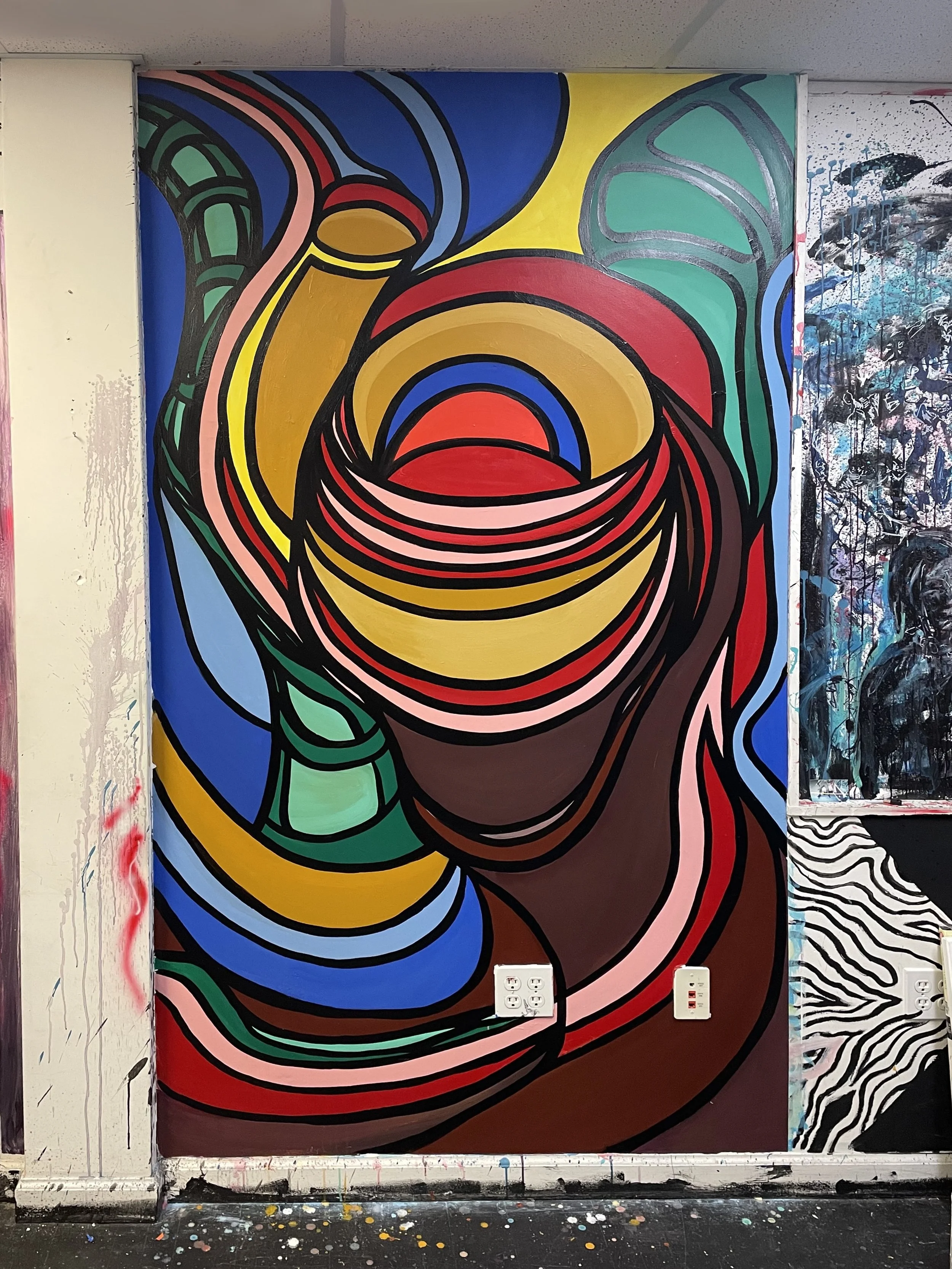 Colorful abstract mural painting with swirling shapes and bold black outlines in red, blue, green, yellow, pink, and brown, on a wall in an art studio or gallery.