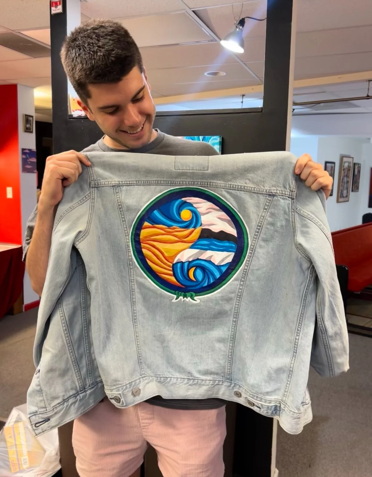 A young man smiling and holding up a light blue denim jacket with a colorful circular embroidered patch depicting a stylized landscape with waves and mountains.