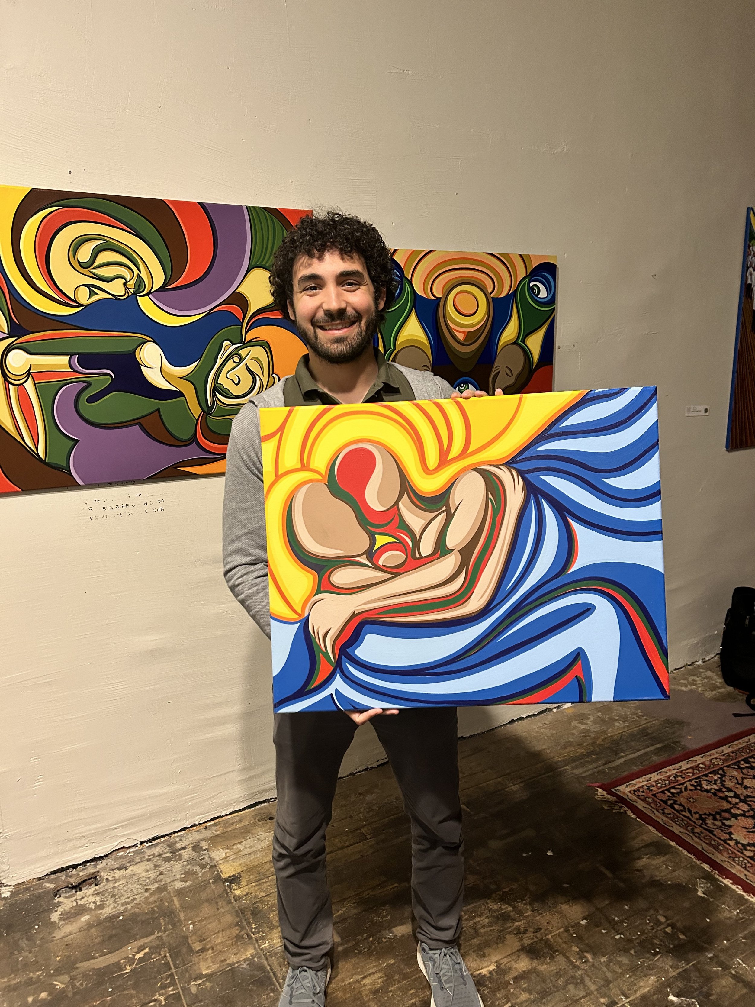 A man holding a colorful abstract painting in an art gallery, with similar artworks visible on the wall behind him.
