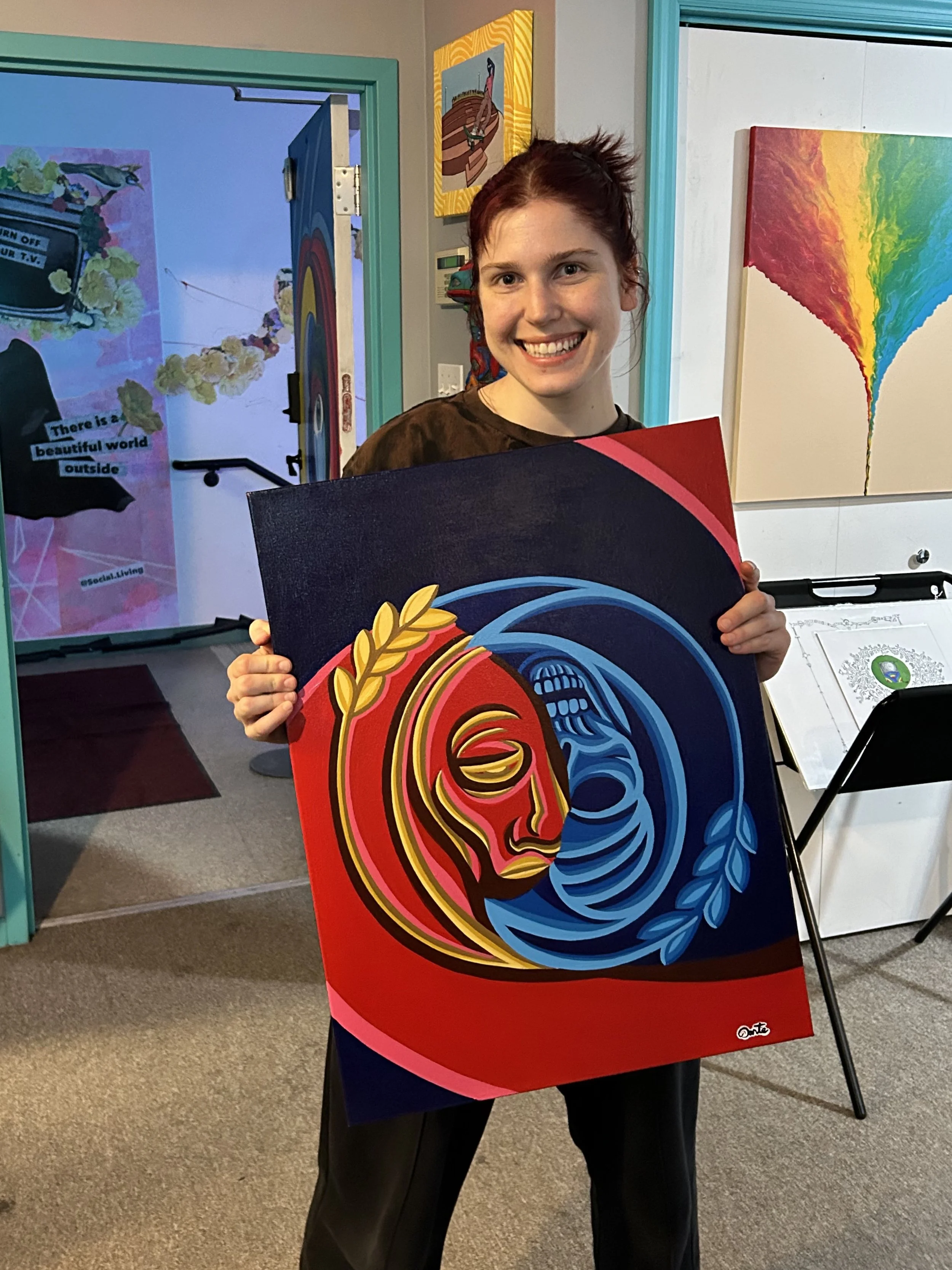 A smiling woman with red hair holds a vibrant, colorful abstract painting featuring a stylized face with red, yellow, and black on the left and blue tones on the right. The background includes an art studio with paintings and a door.