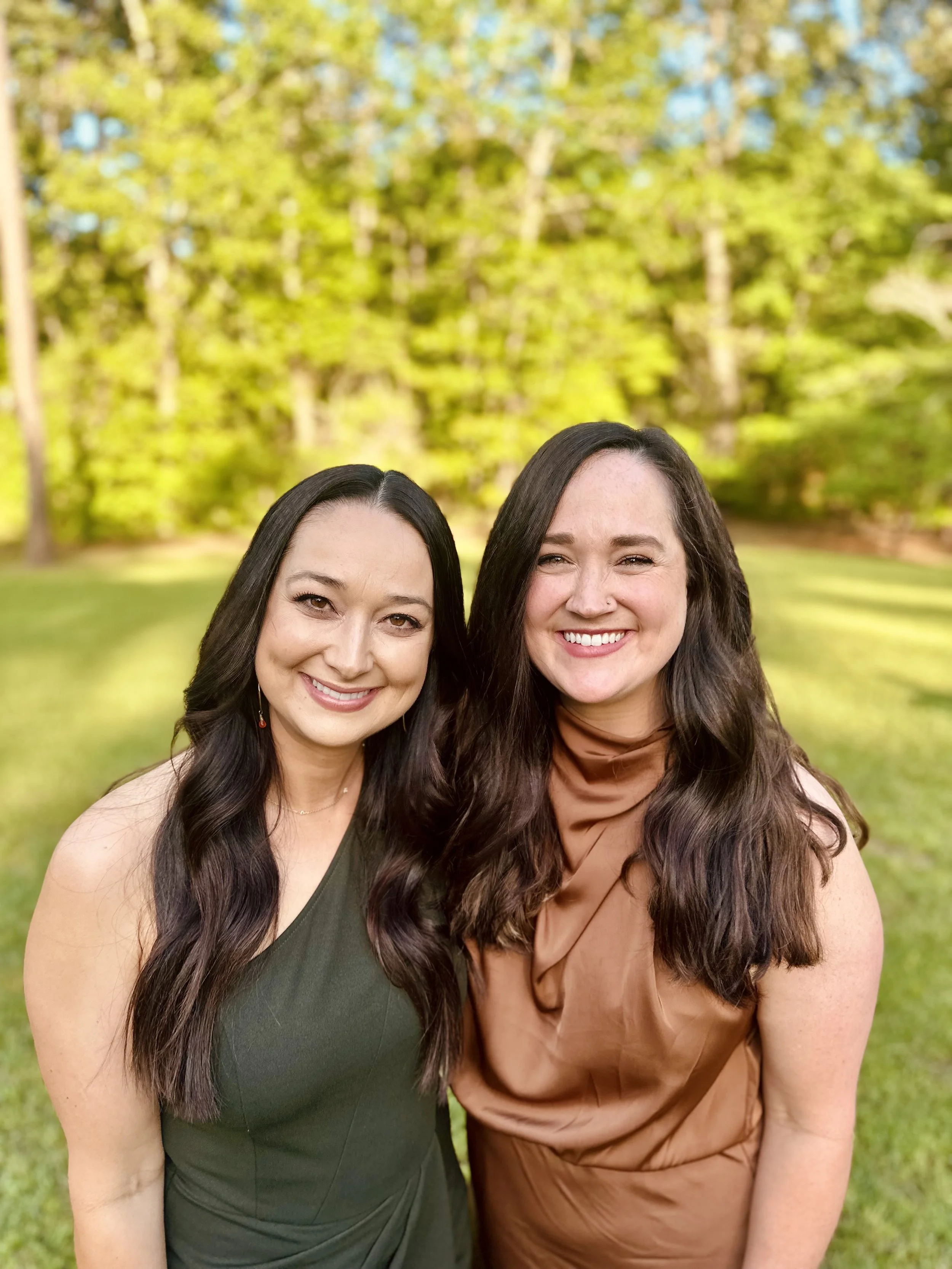 Two women smiling outdoors in a green park with trees in the background.