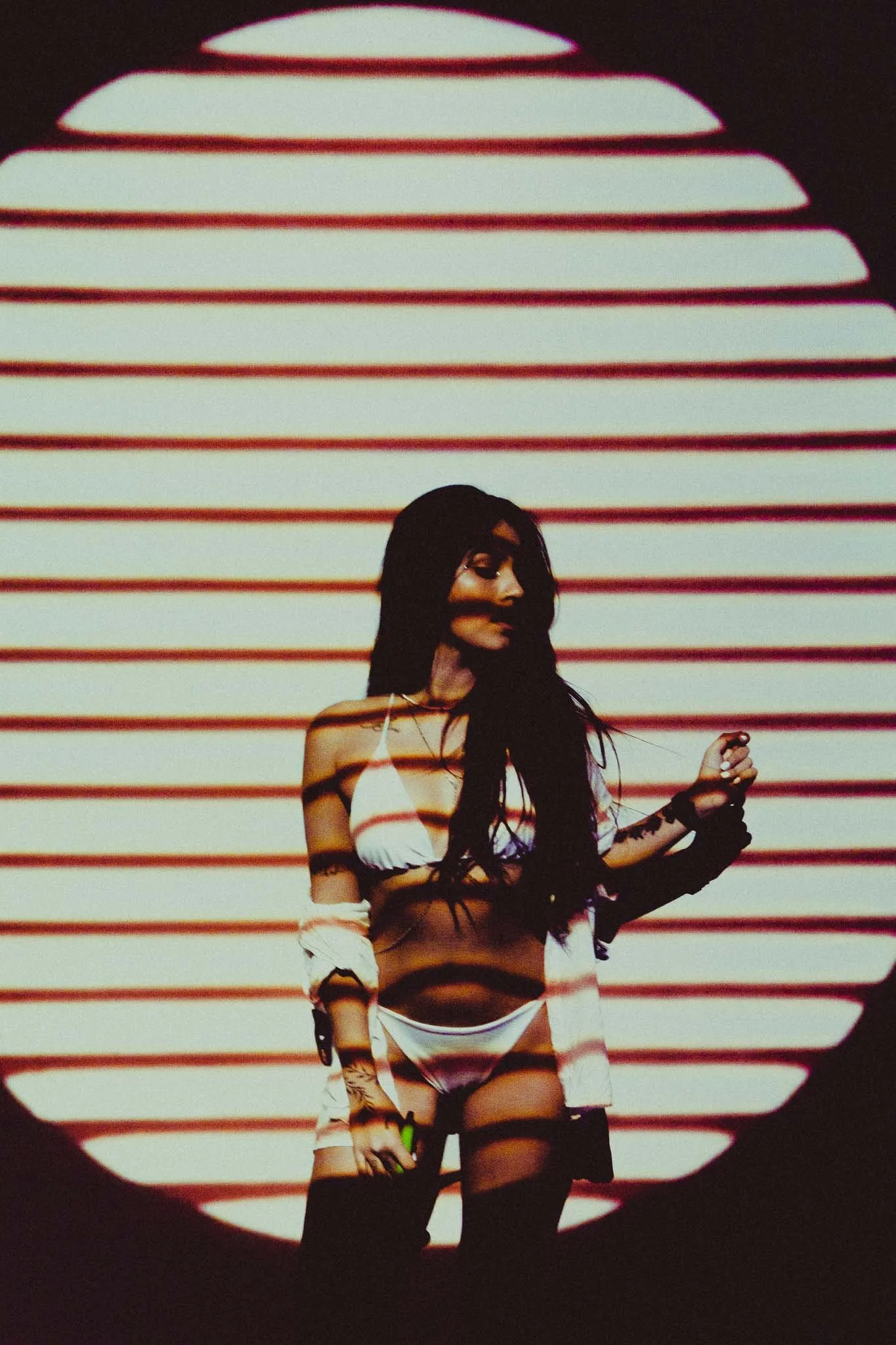 A woman with long dark hair in a bikini and a light jacket stands in front of a striped projected background, with alternating dark and light lines casting shadows and light across her body.