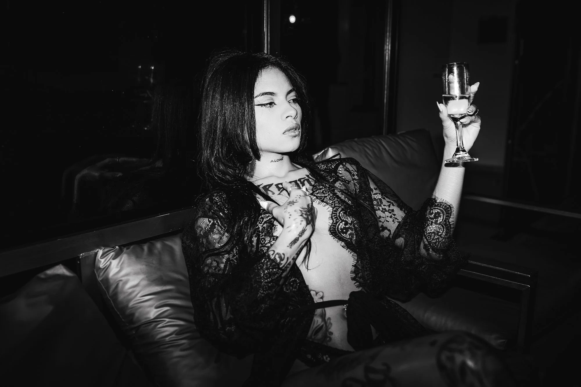 A woman with dark hair and tattoos, wearing lace clothing, is sitting on a sofa holding a glass of champagne, in a dimly lit room.