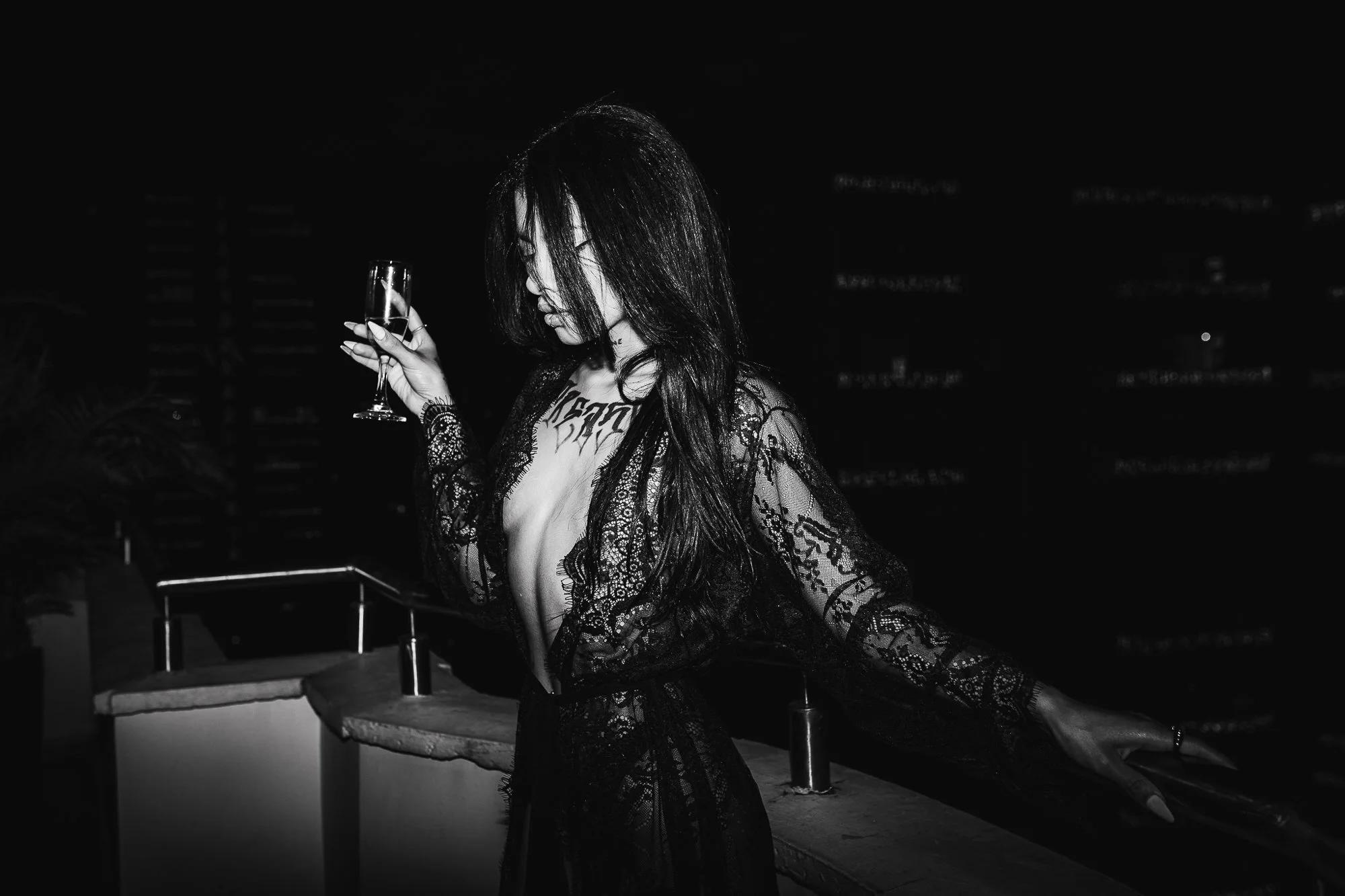 A woman in a black lace robe holds a glass of champagne on a rooftop at night.