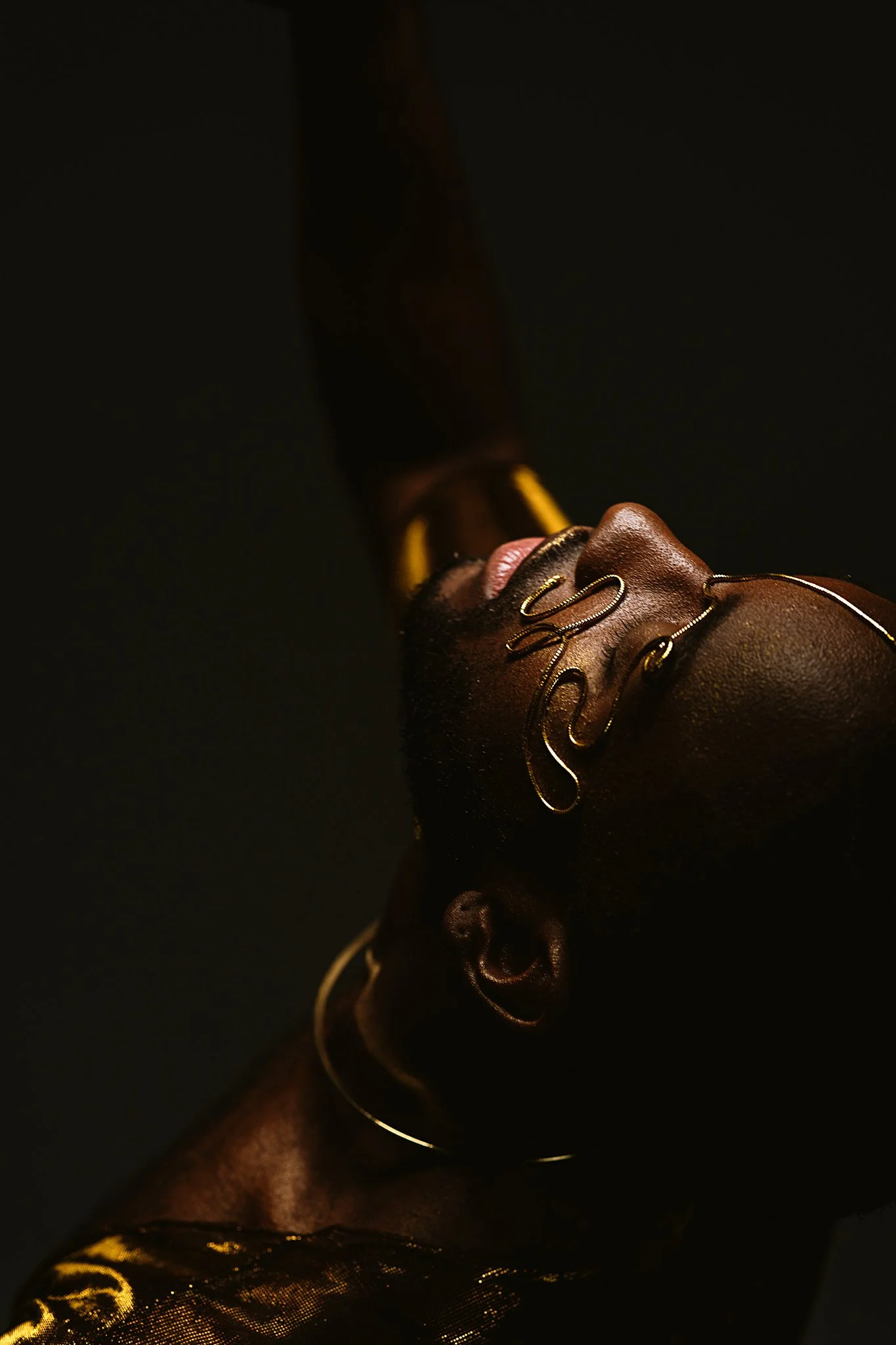 Close-up of a Black woman with gold jewelry, lying down with her face turned upward against a black background.