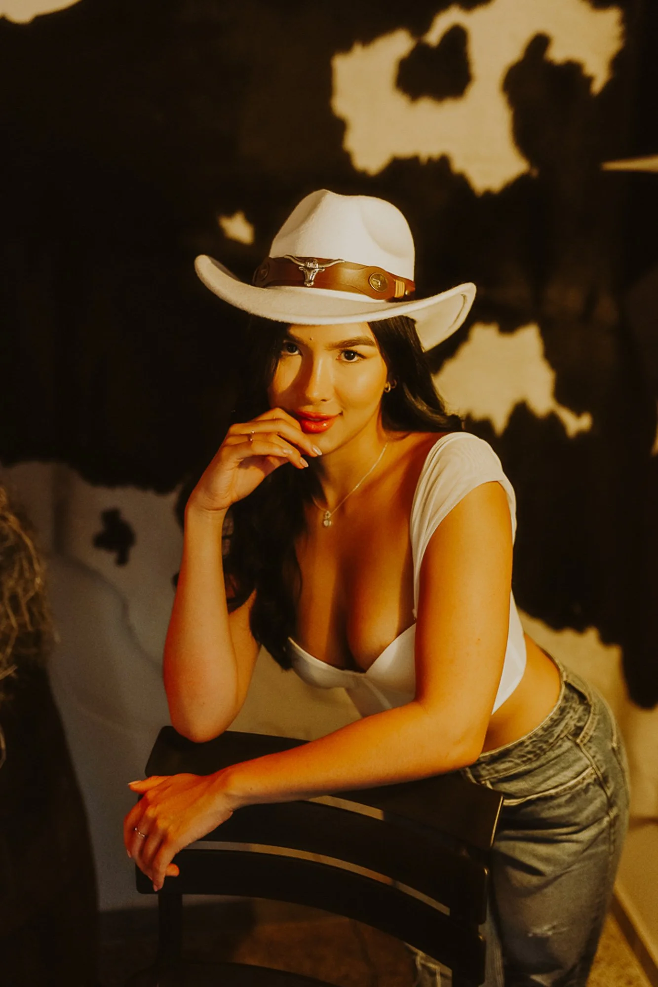 A woman wearing a white cowboy hat, a white top with a deep neckline, and ripped jeans, leaning on a black chair, with a dark abstract wall behind her.