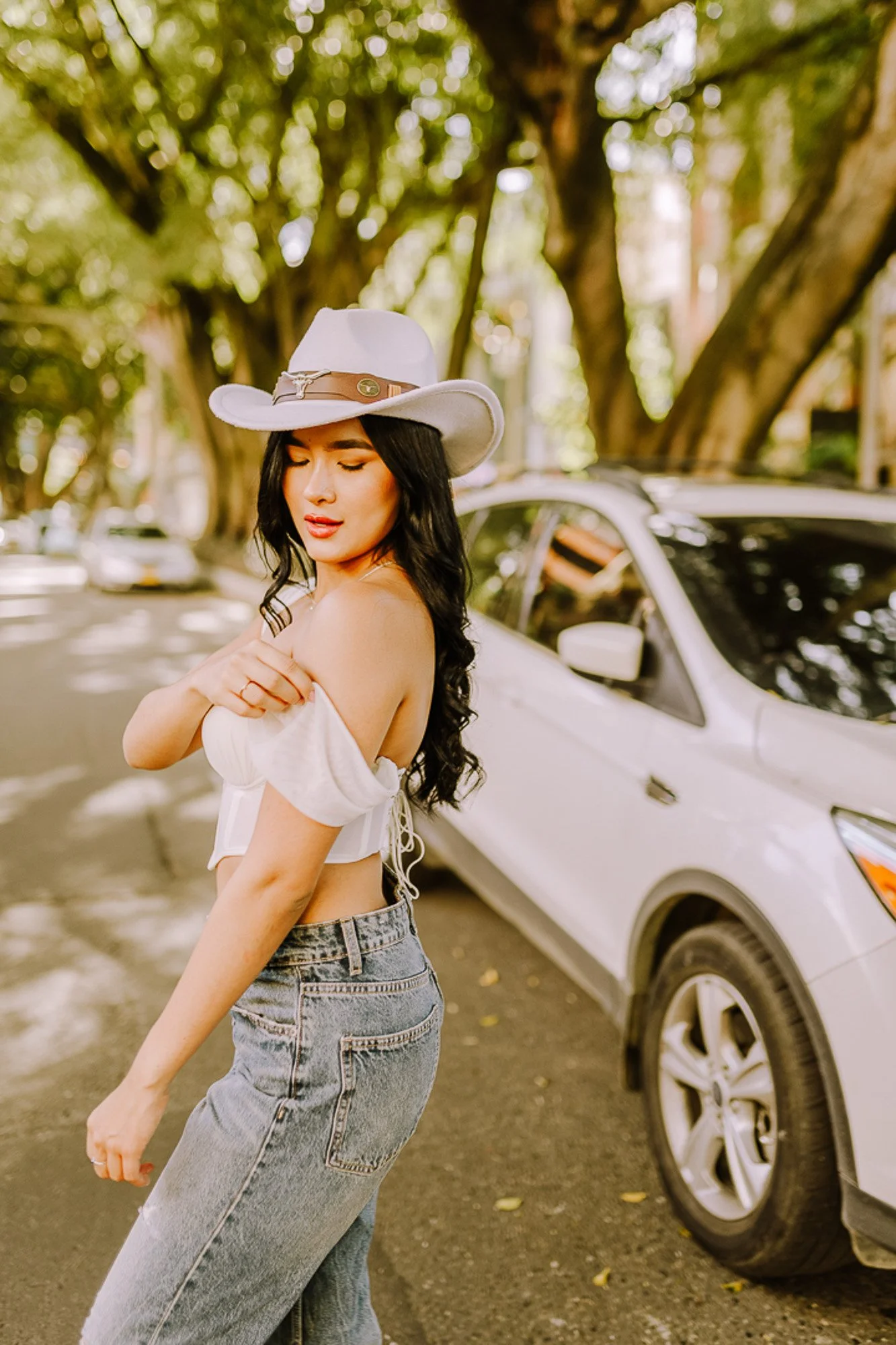 A woman standing next to a white SUV on a tree-lined street, wearing a white off-the-shoulder top, high-waisted jeans, and a white cowboy hat with a brown band.