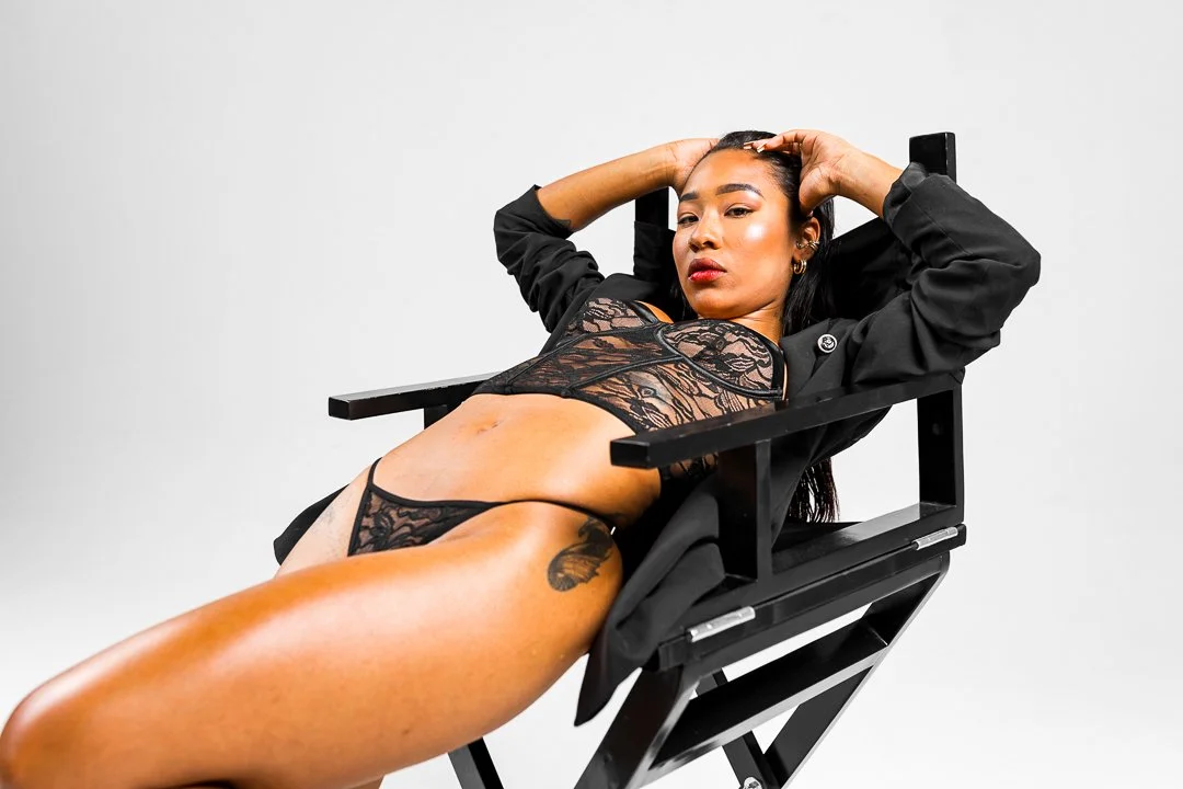 A woman in black lace lingerie and a black blazer reclining on a director's chair against a plain white background.
