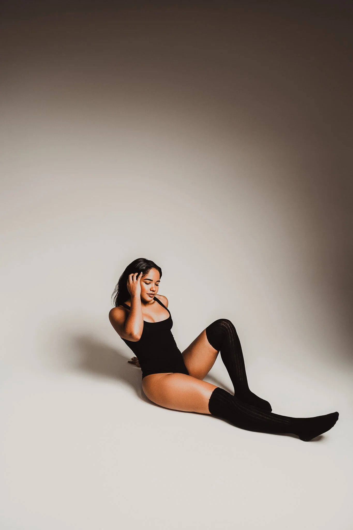 A young woman with dark hair sitting on the floor, wearing black thigh-high socks and a black one-piece bodysuit, against a plain light background.