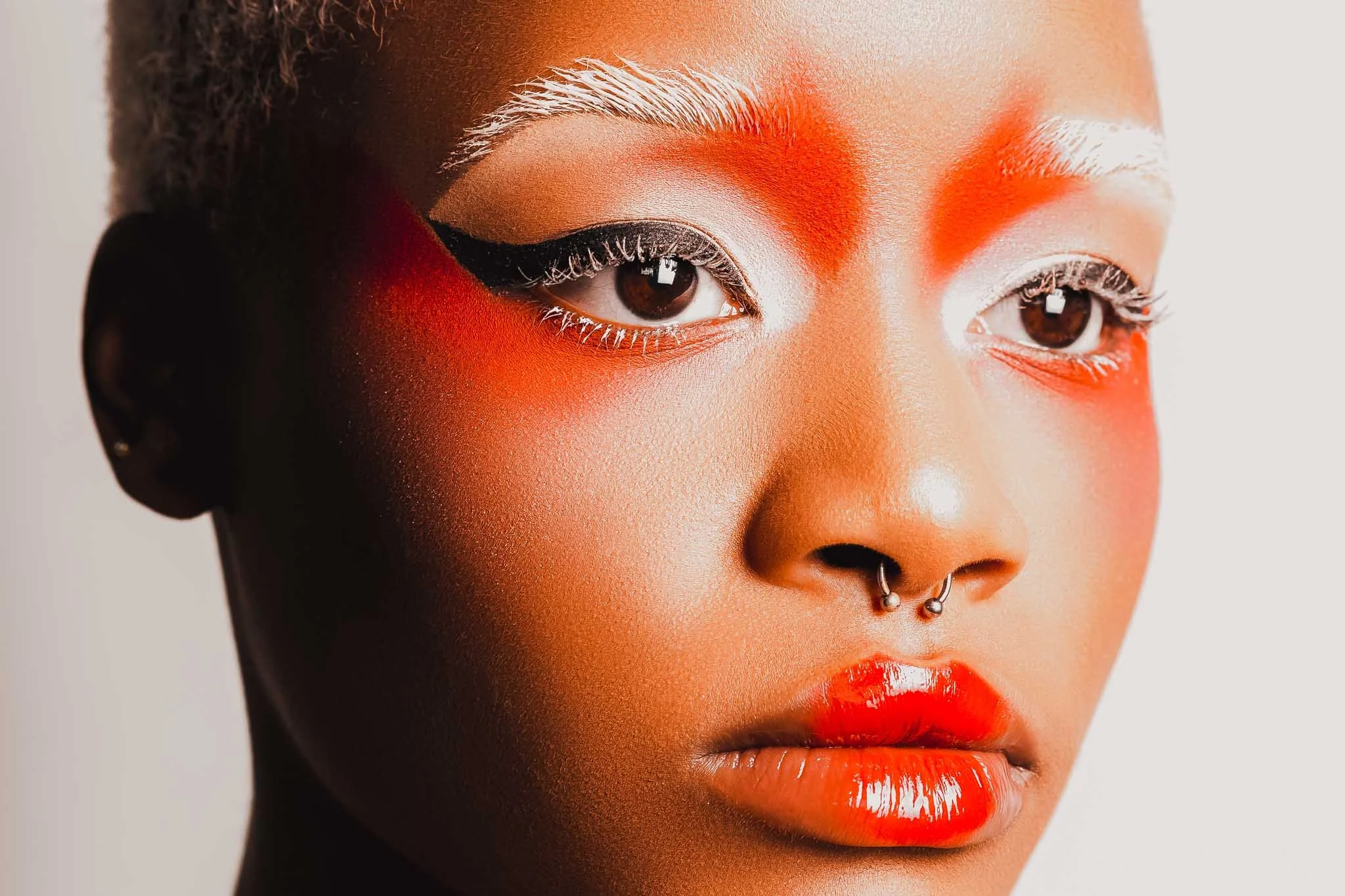 Close-up of a woman with bold makeup featuring white, black, and red eyeshadow, white eyebrows, white eyeliner, and bright red lipstick. She has a septum piercing and dark brown eyes.