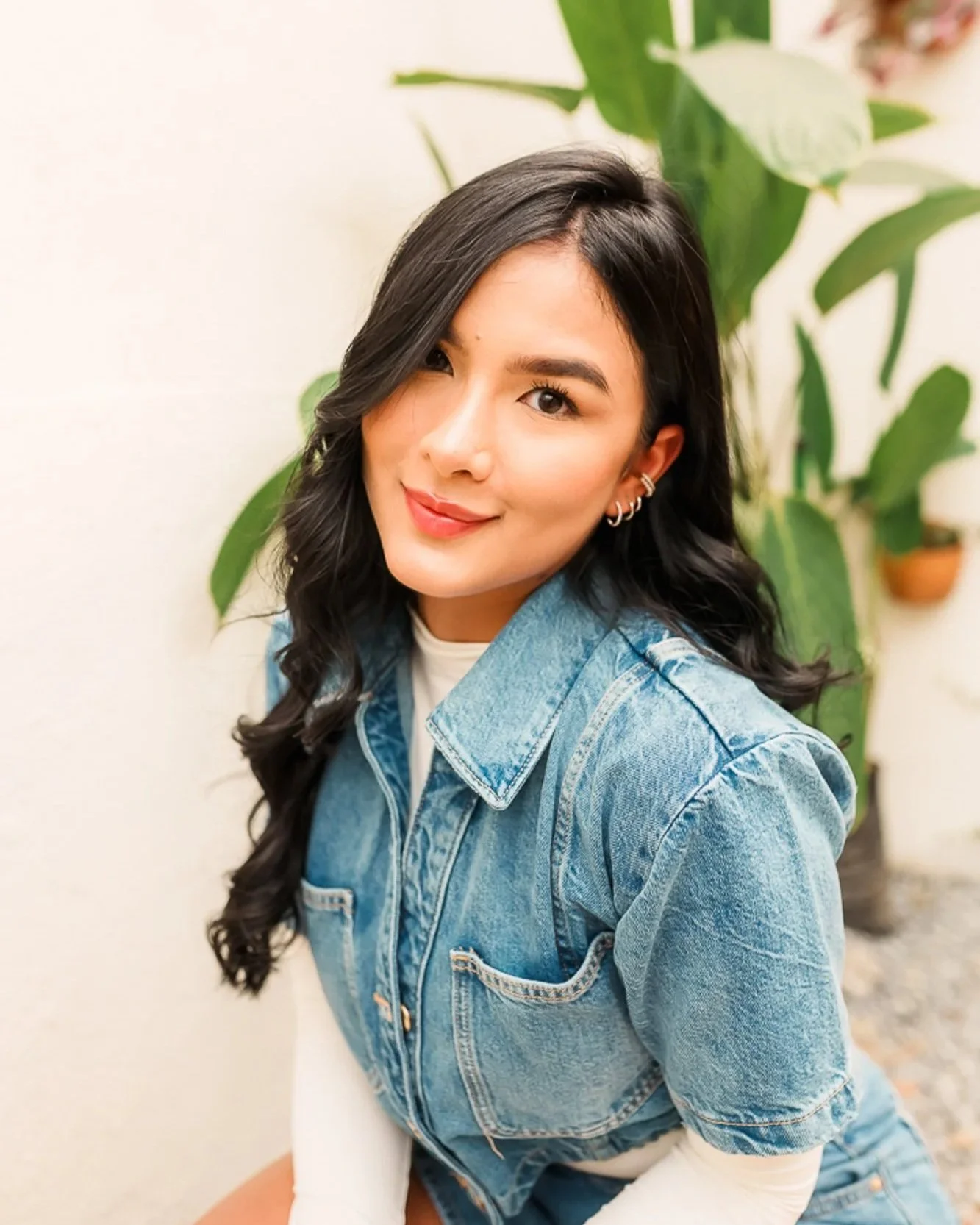 A young woman with black, wavy hair winking and smiling, wearing a denim jacket and earrings, standing in front of a leafy green plant.