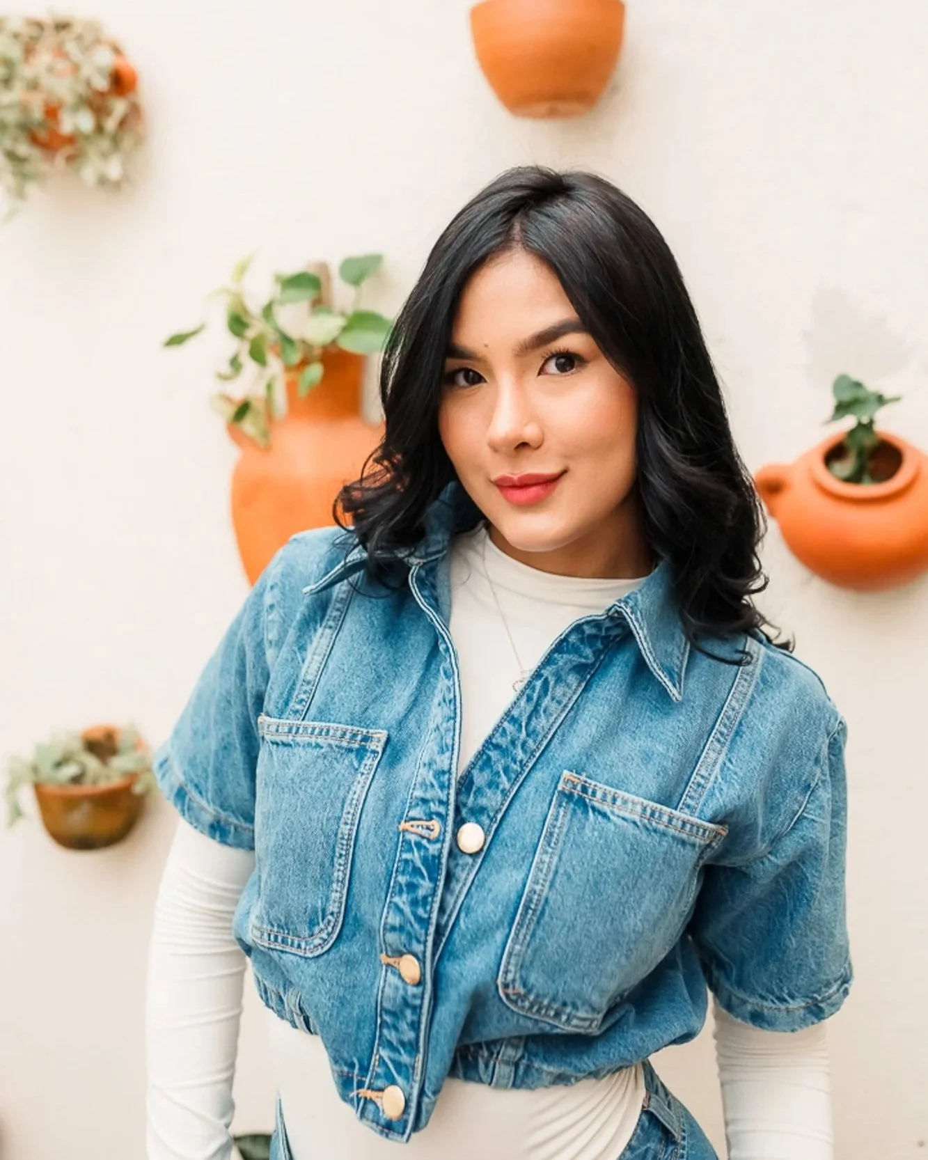 A woman with black wavy hair wearing a white shirt and denim jacket standing in front of a wall decorated with hanging terracotta pots with green plants.
