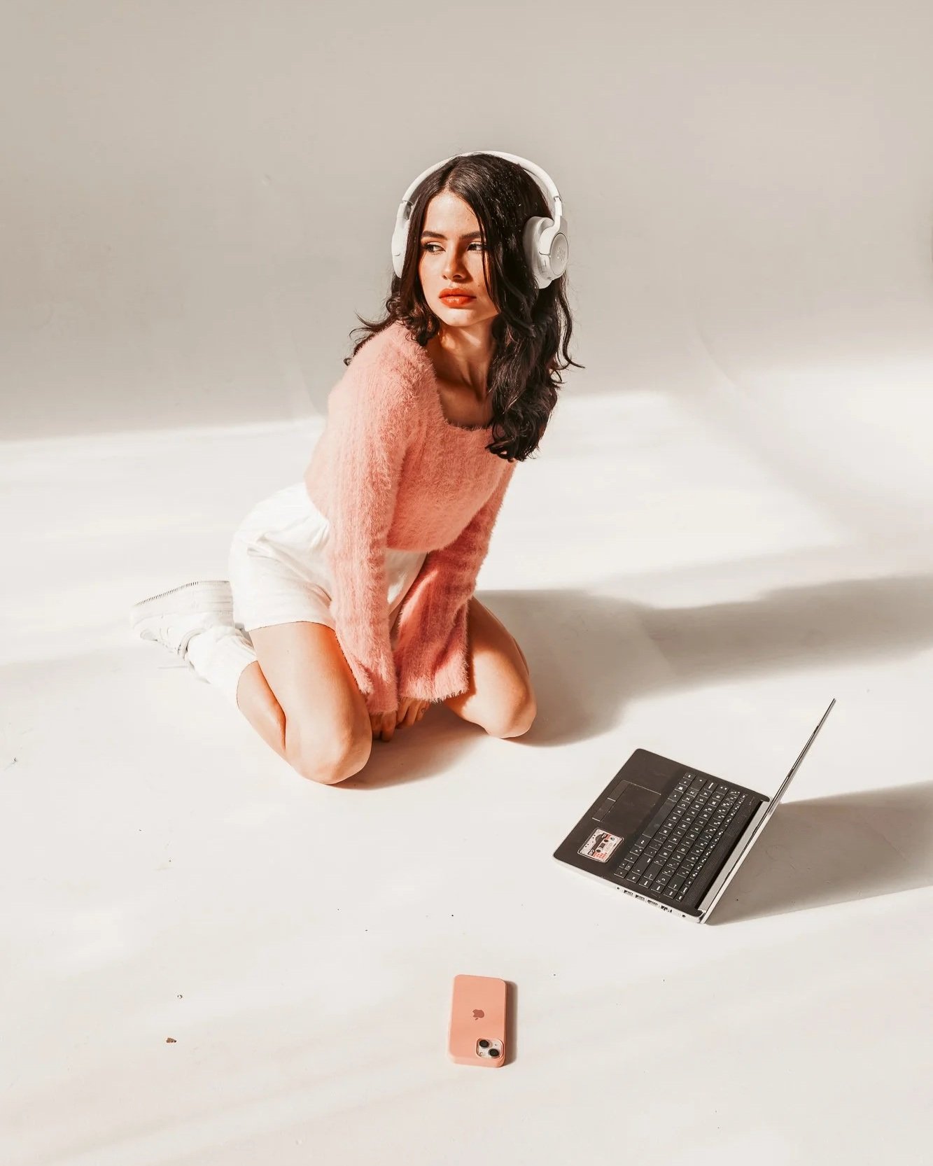A woman in a pink fuzzy sweater and white shorts kneels on the floor, wearing large white headphones, with a laptop and pink phone nearby on a plain white background.