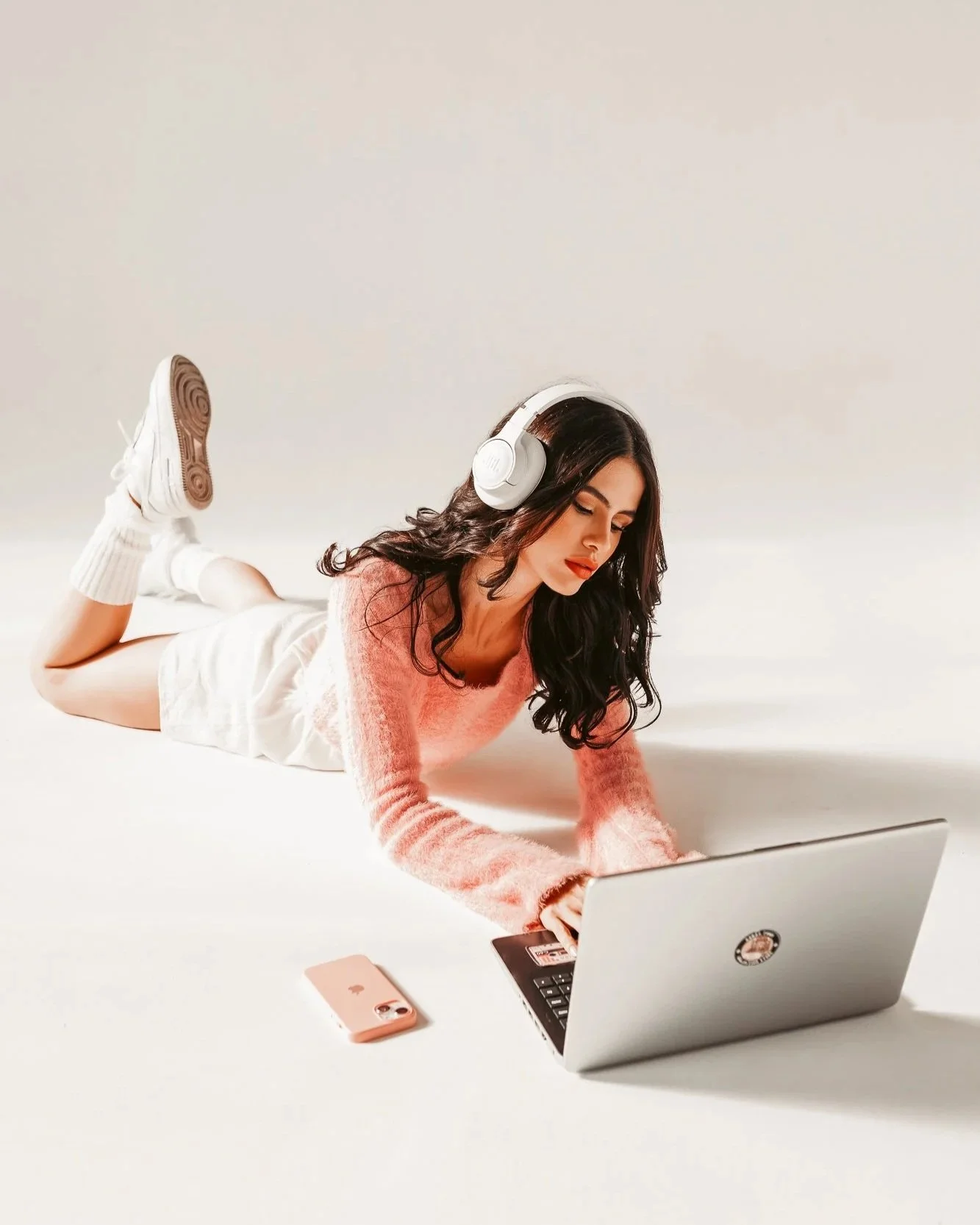 Woman lying on the floor using a laptop, wearing headphones, with a smartphone nearby, against a plain background.