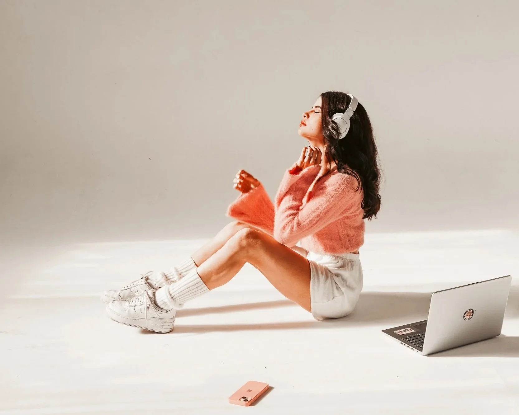 A young woman with long wavy dark hair sitting on the floor with her legs extended, wearing a pink sweater, white shorts, white socks, and sneakers. She is wearing large headphones, leaning back with her eyes closed, enjoying music. A laptop is on th