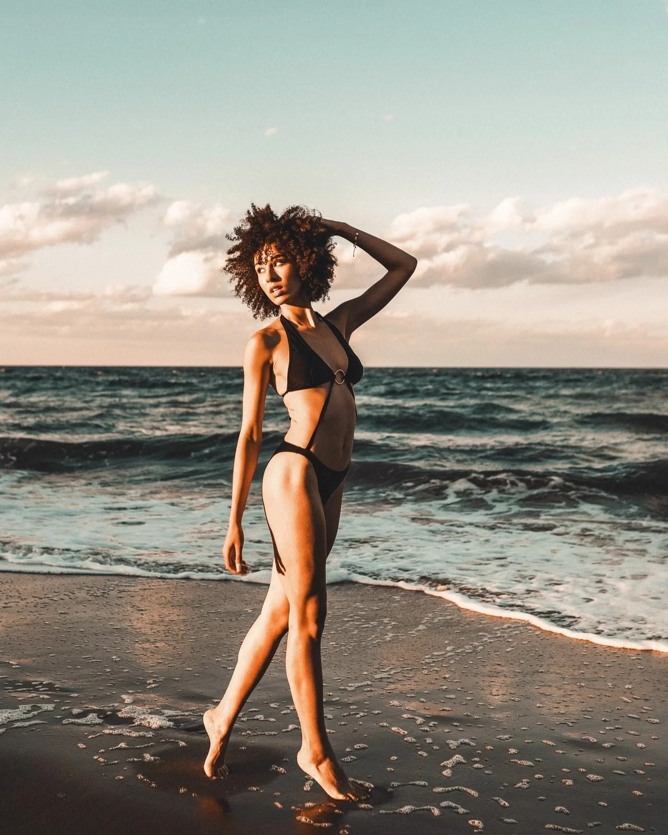 Woman in black bikini walking on the beach with ocean waves and cloudy sky in the background