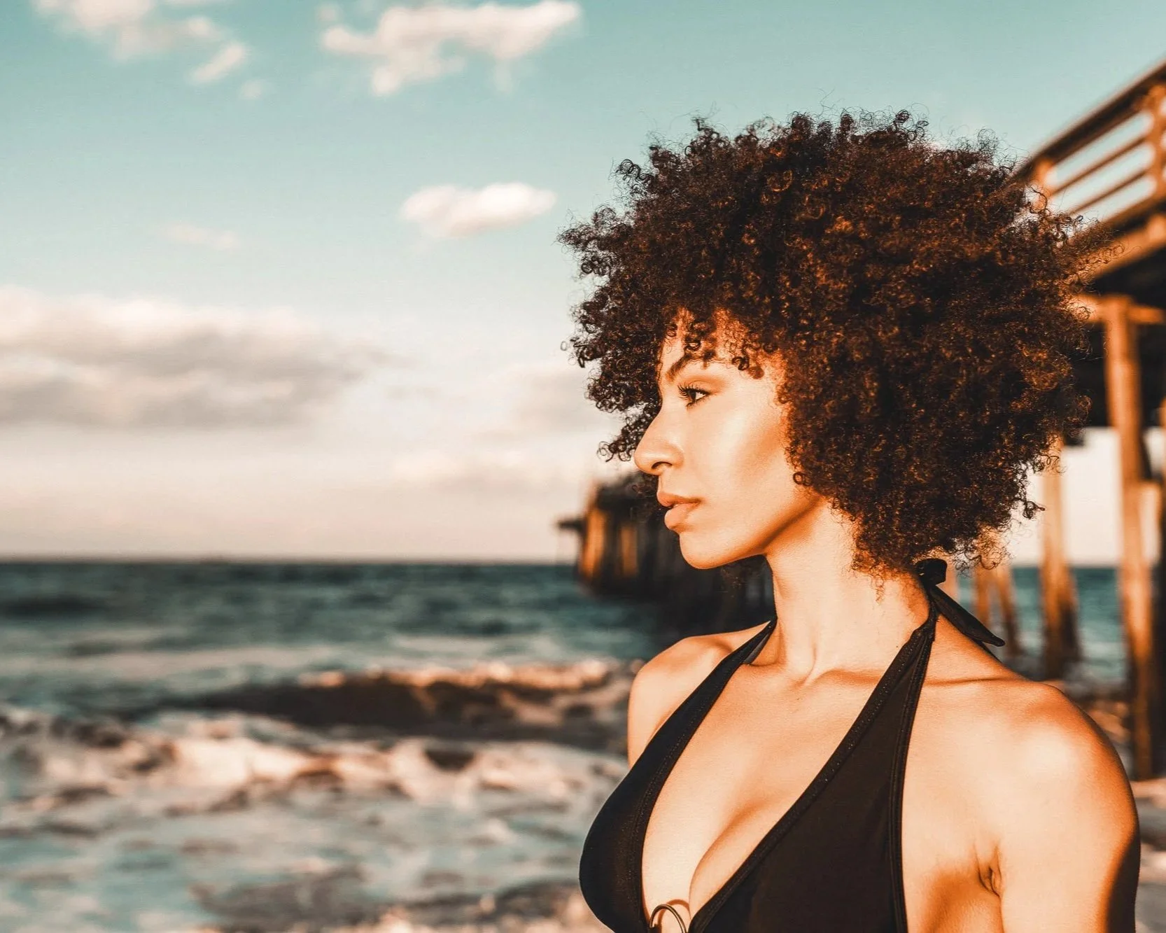 A woman with curly hair wearing a black bikini top standing on a beach during sunset, with pier structures visible in the background.