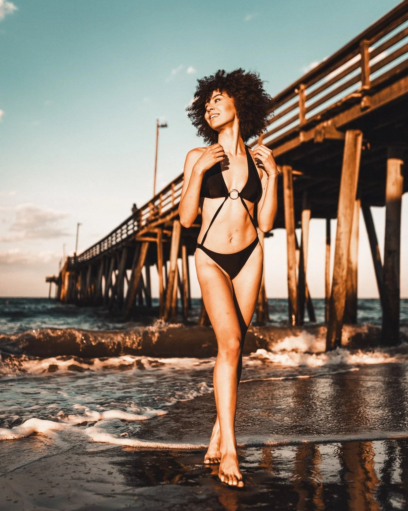 A woman in a black bikini walking on a beach near a pier at sunset.