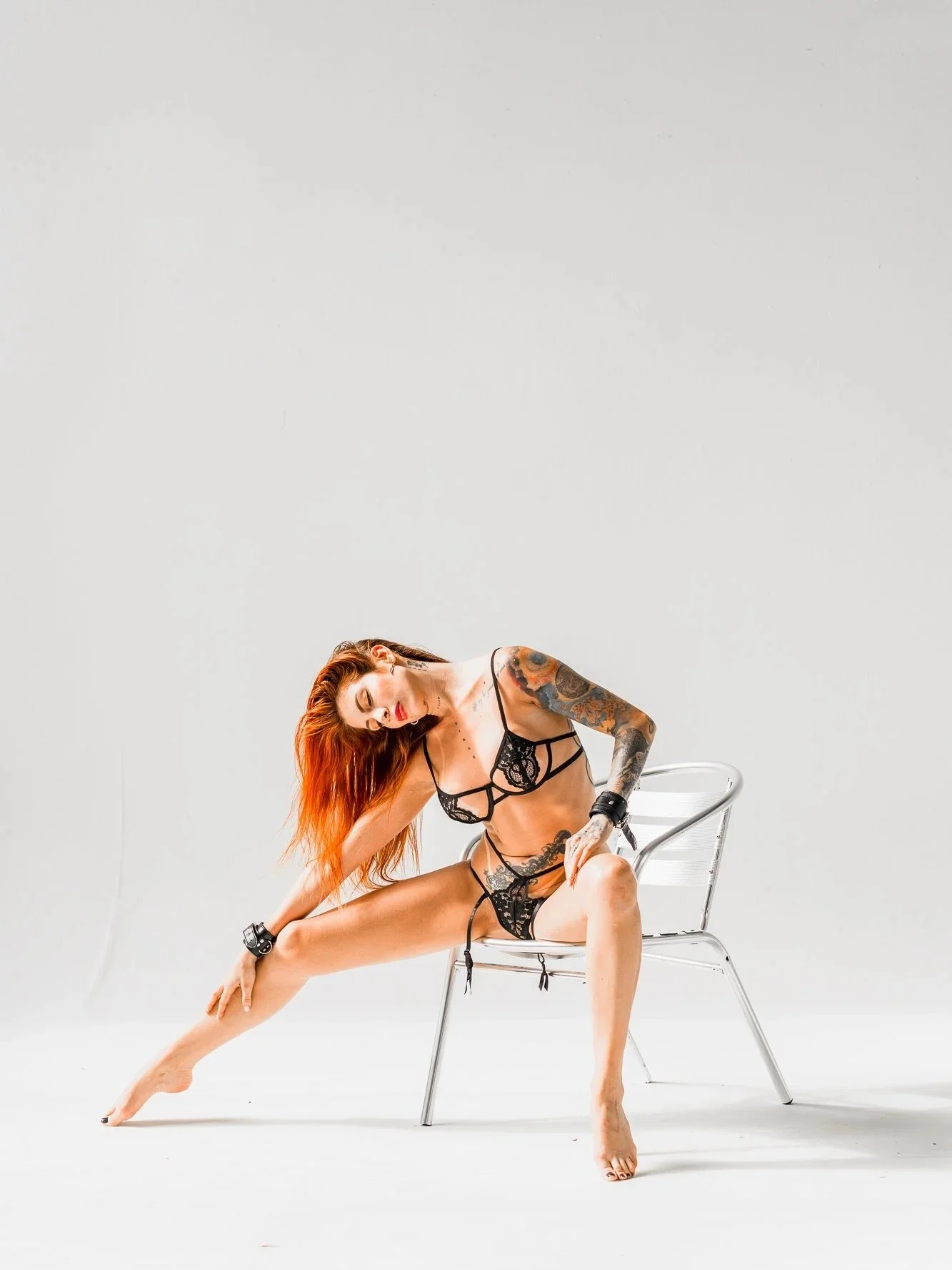 A woman with red hair, tattoos, wearing black lace lingerie, poses sitting on a silver chair with her legs spread, leaning to one side with a gray background.