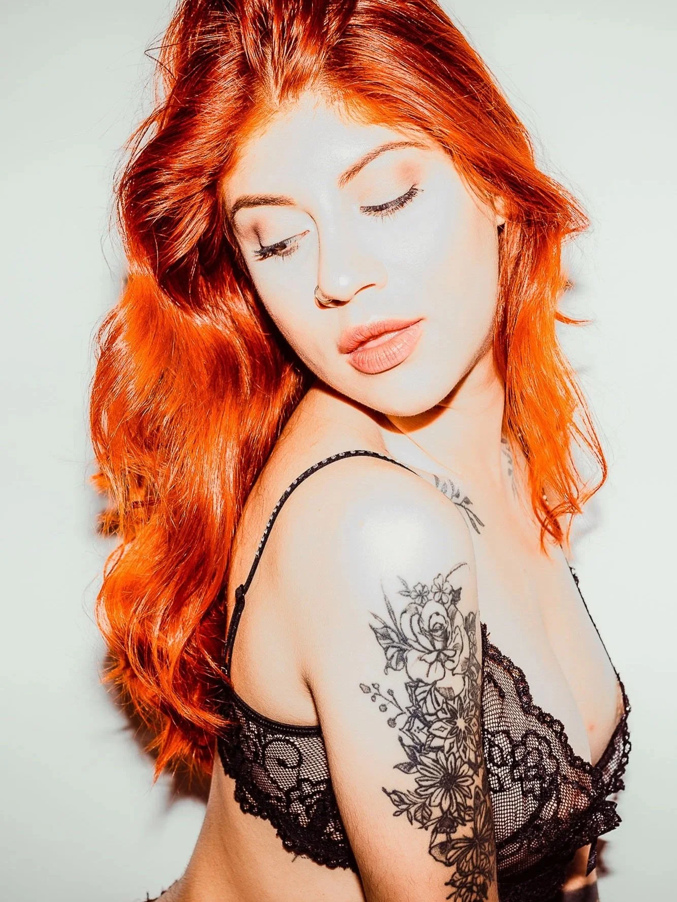 A woman with long, wavy red hair wearing black lace lingerie, showing a floral tattoo on her upper arm, with her eyes closed and head slightly tilted.