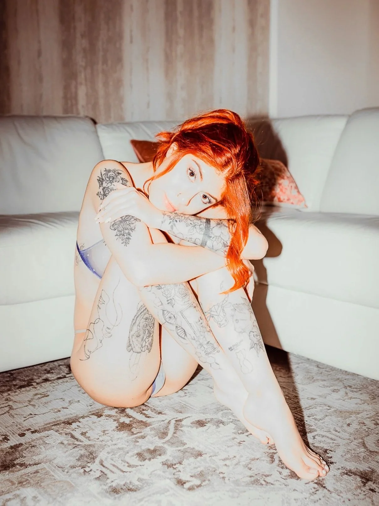 A woman with red hair and tattoos sitting on the floor in front of a white sofa, hugging her knees, with soft lighting and a textured wall behind her.