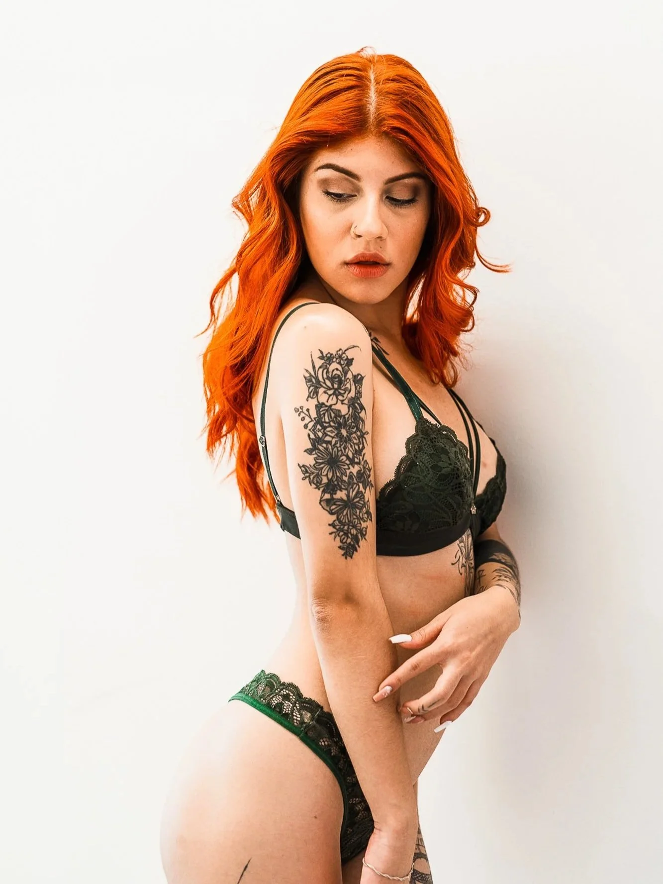 A woman with long, wavy red hair and tattoos on her arm and side, wearing black lace lingerie, standing against a plain white background.