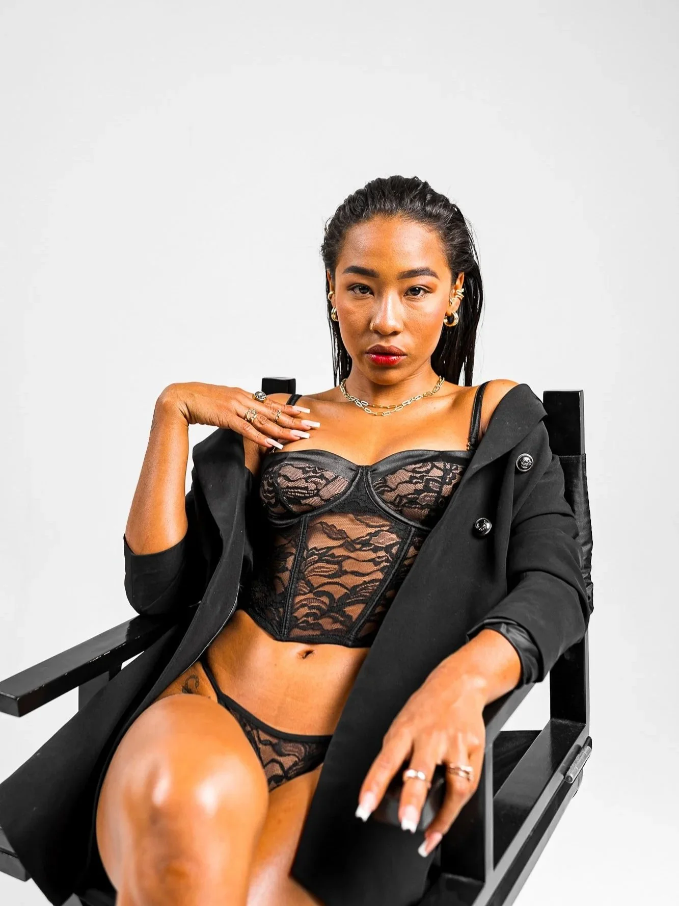 A woman sitting in a black chair, wearing black lace lingerie and a black jacket, with jewelry including rings, earrings, and necklaces, posing against a plain white background.