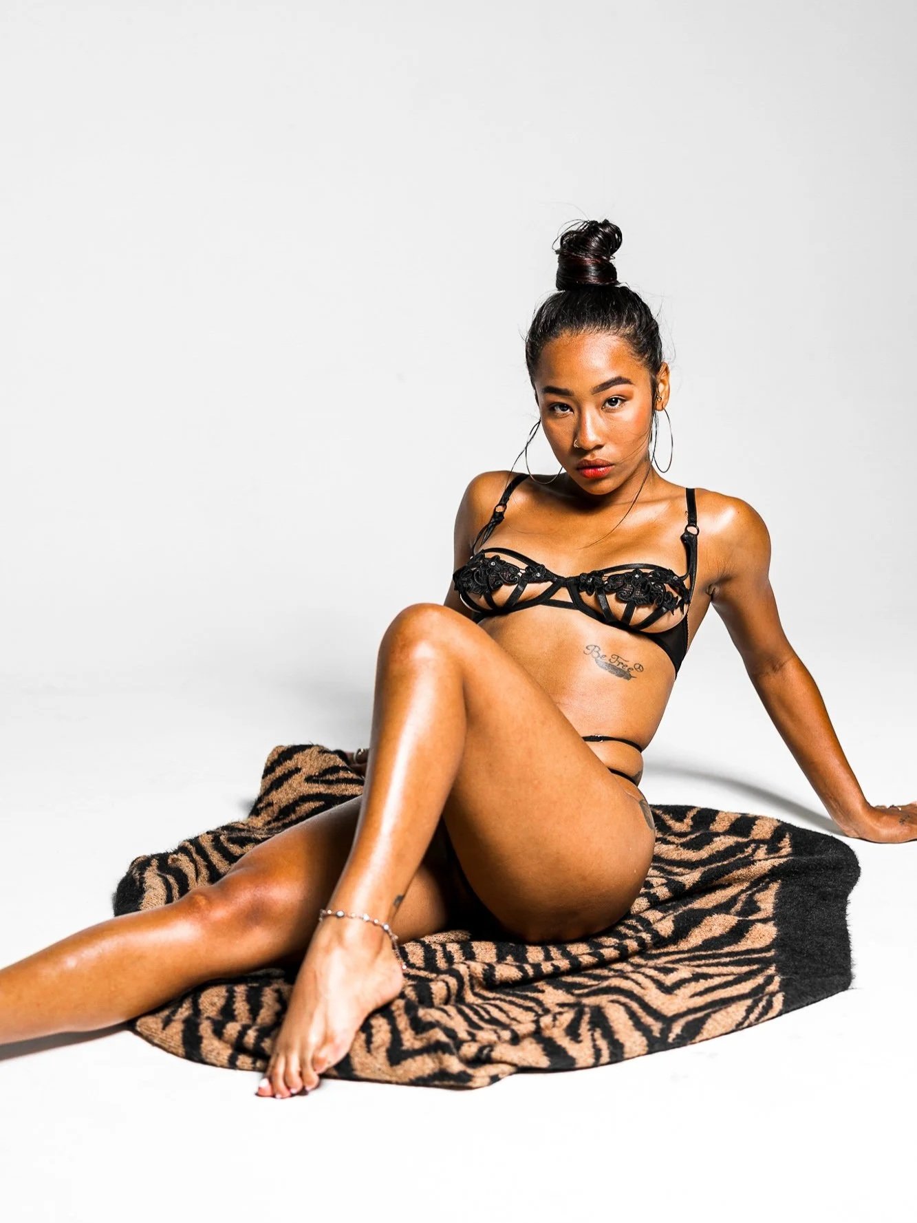 A woman with dark hair in a top knot, wearing black lingerie with strappy details, sitting on a tiger-print blanket on the floor against a white background. She has hoop earrings and tattoos on her side and ankle.