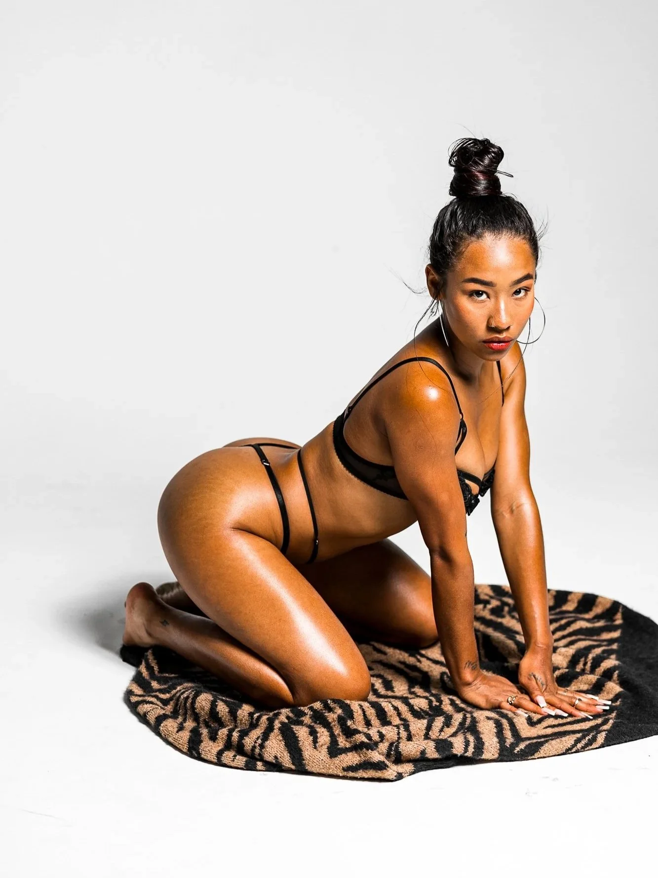 Woman with dark hair styled in a high bun, kneeling on a tiger print blanket, wearing black lingerie, posing on a white background.