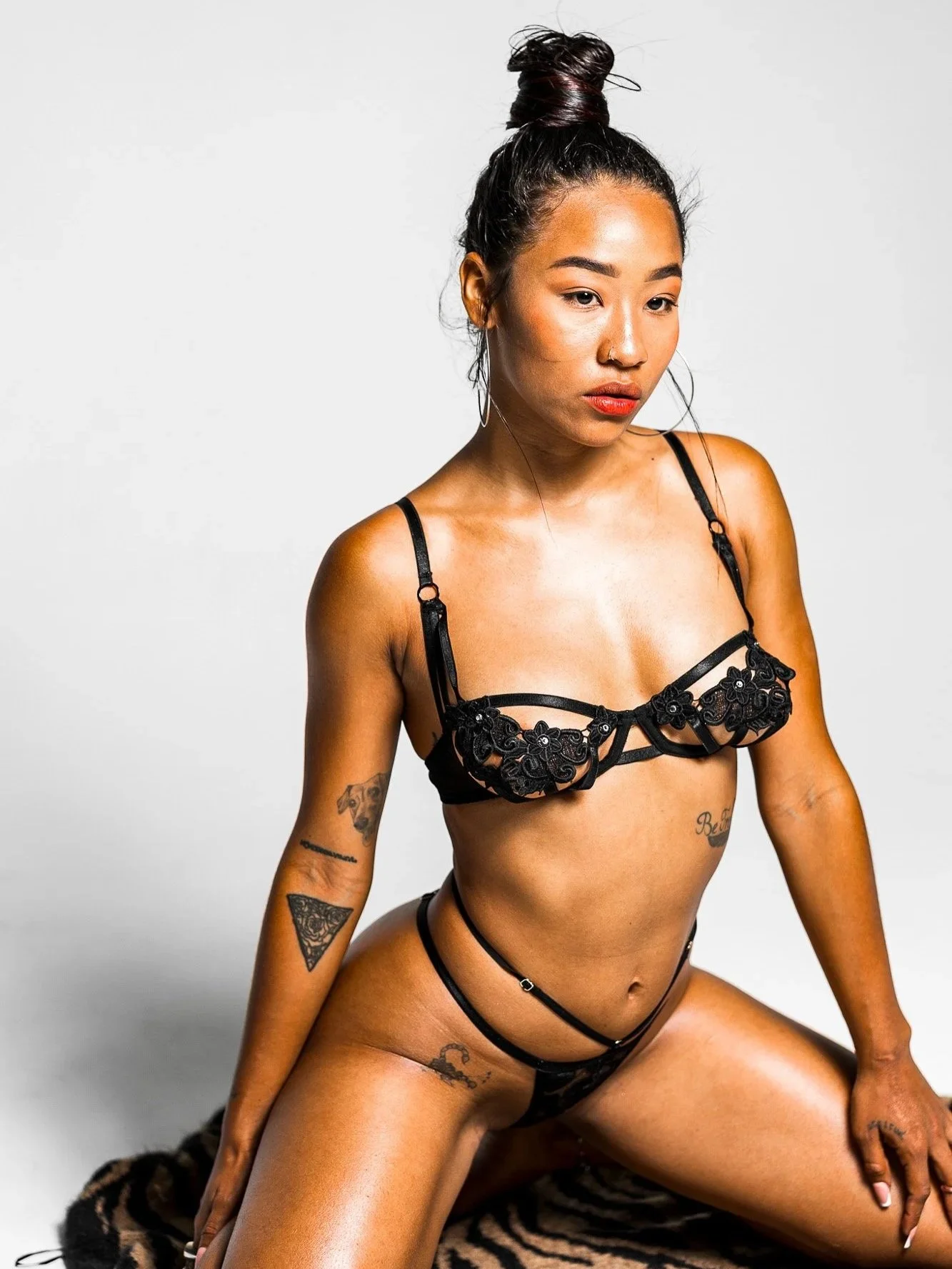 A woman with tattoos poses on a zebra print rug, wearing black lingerie with floral lace details, against a plain white background.
