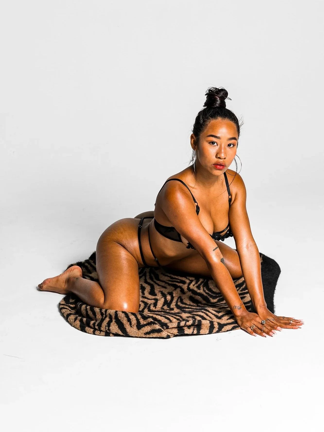 A woman with dark hair in a high bun, dressed in black lingerie, kneeling on a tiger-print rug against a white background, looking directly at the camera.