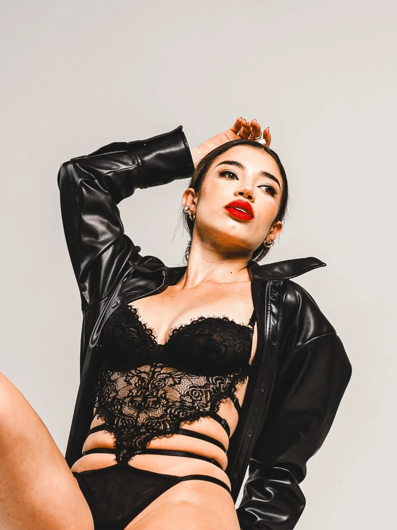 A woman with dark hair, wearing a black lace bralette, matching panties, and a black leather jacket, poses against a plain white background.