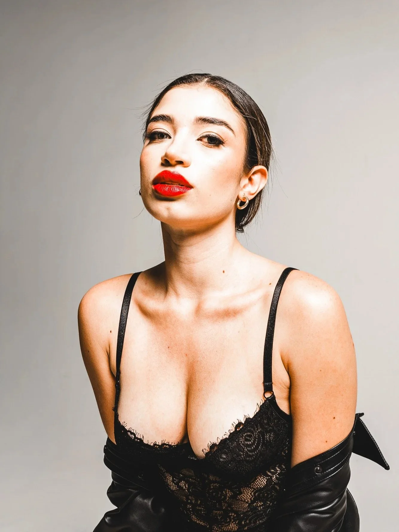 A woman with dark hair, red lipstick, wearing earrings, a black lace top, and a black leather jacket draped over her shoulders, posing confidently against a plain background.