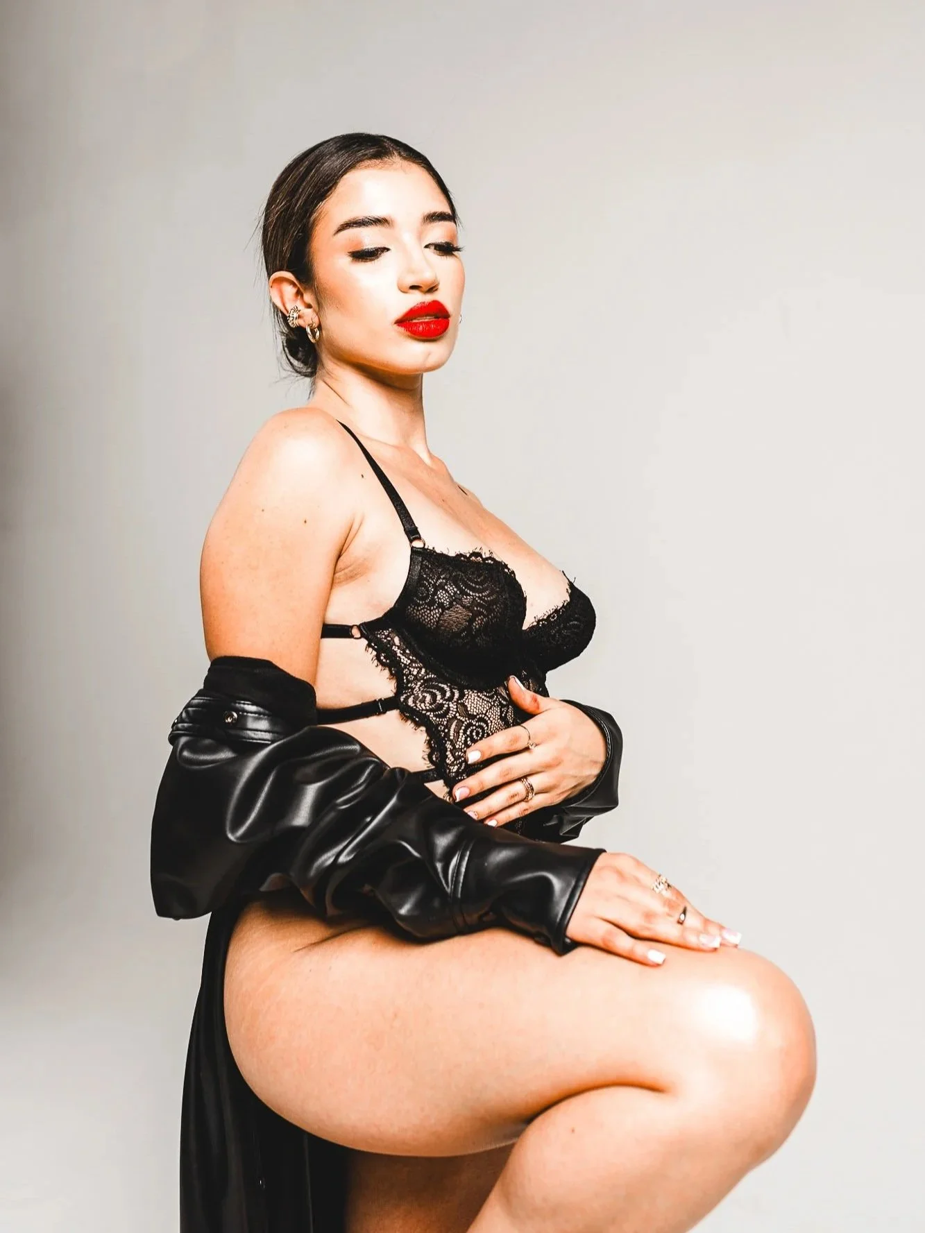 A woman with dark hair and red lipstick wearing black lace lingerie and a black leather jacket draped over her arms, kneeling with one knee raised against a plain background.