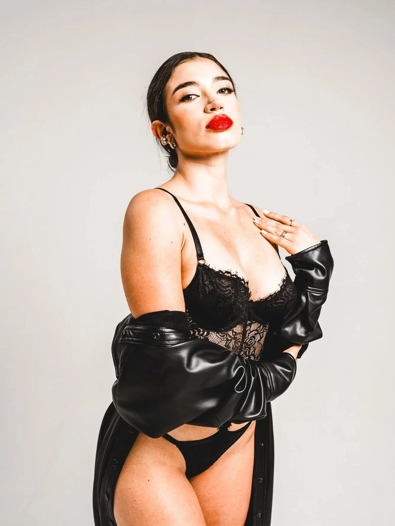 A woman with dark hair and bold red lipstick posing in black lingerie and a black leather jacket against a plain background.