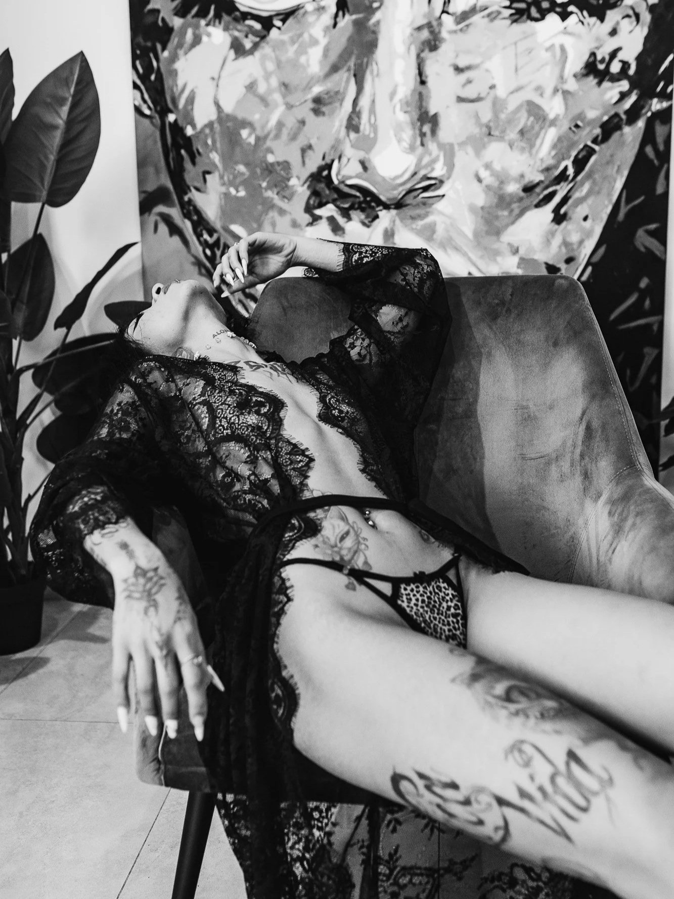 A woman with tattoos and dark hair reclining on a sofa wearing black lace lingerie and a sheer lace robe, with a large abstract painting behind her and a potted plant to her left.