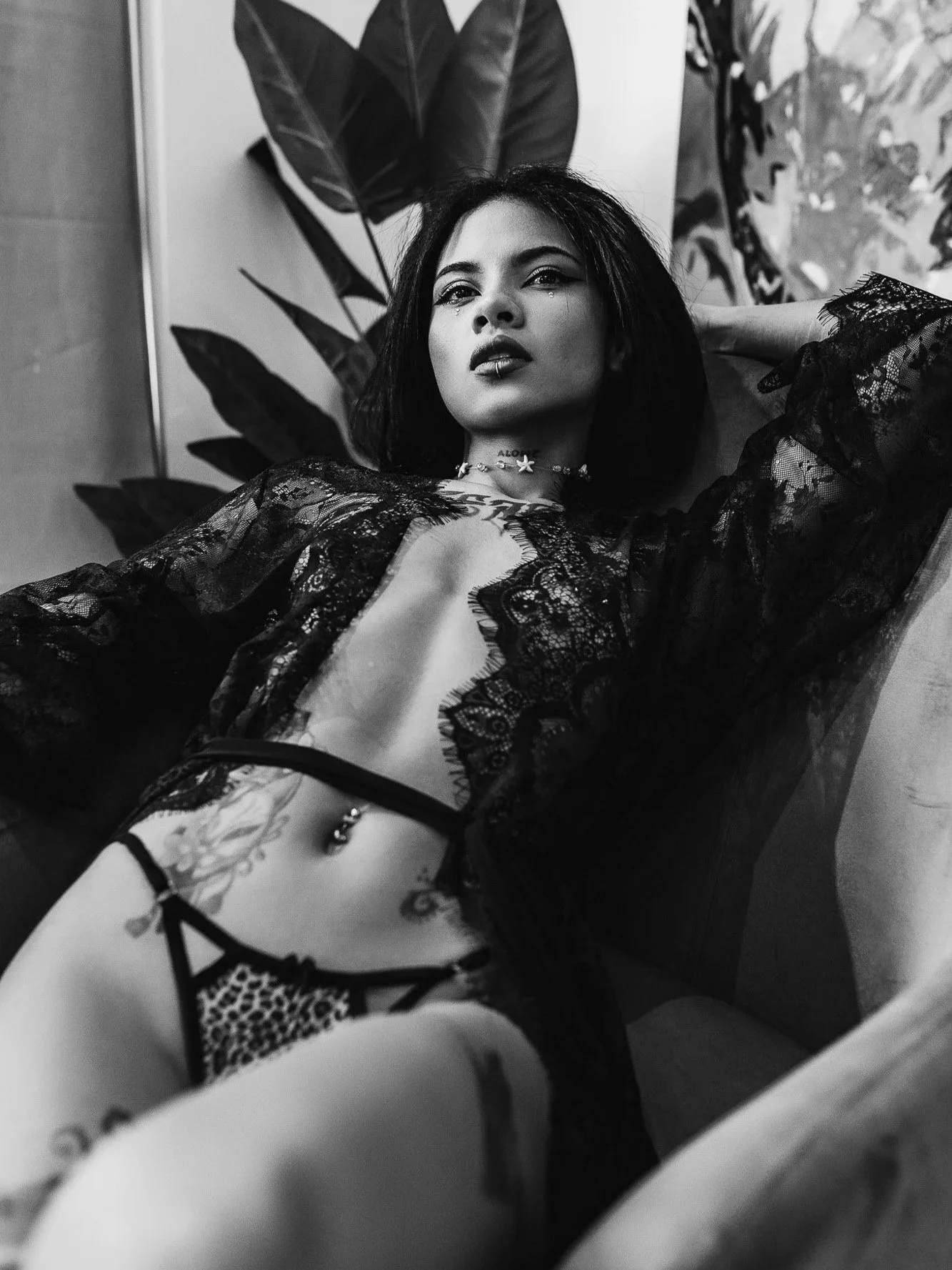 A woman with dark hair reclining on a chair or bed, wearing lace lingerie and jewelry, with plants in the background.
