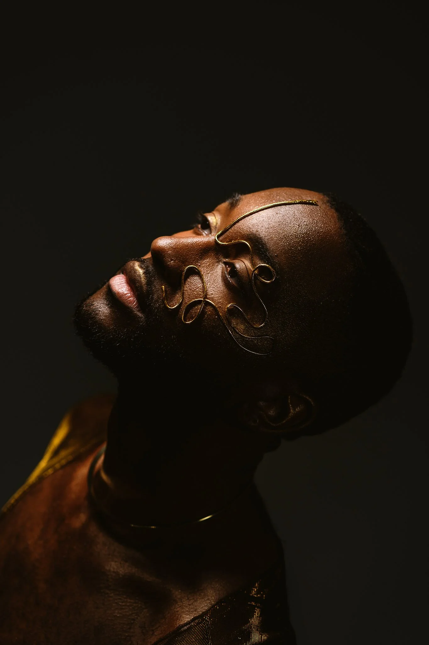 Portrait of a Black person with short hair, wearing artistic gold wire jewelry on face and neck, against a black background.