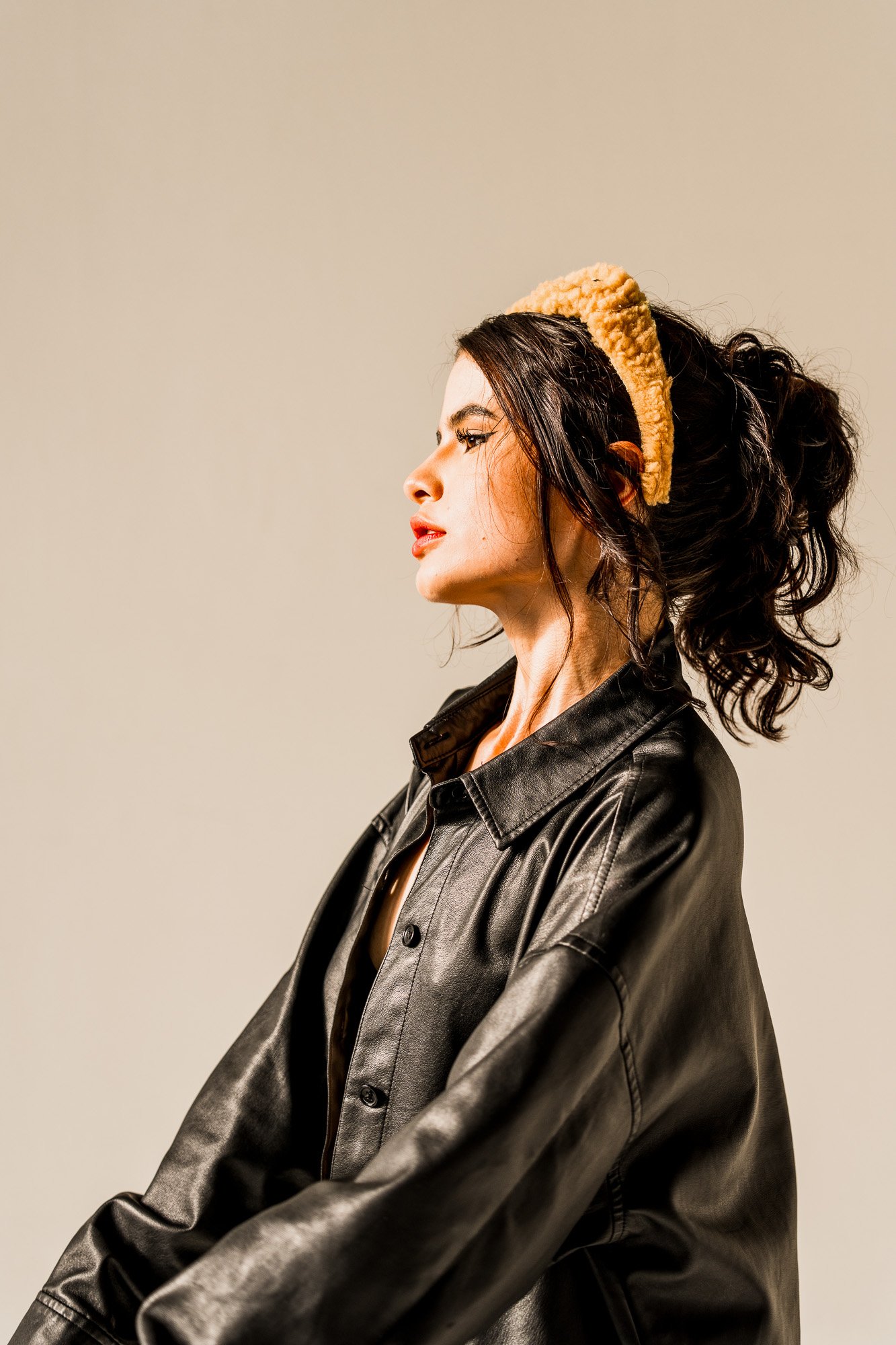 Profile portrait of a woman in a black leather jacket and textured headband, lit by warm studio light against a neutral background.